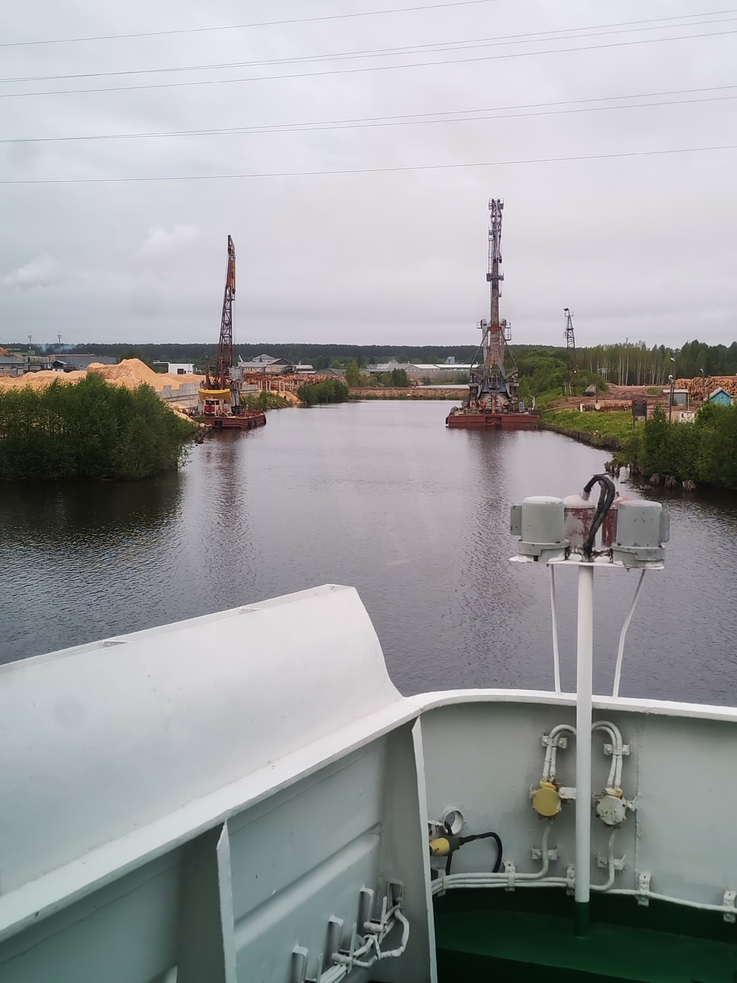The image depicts a view from a ship sailing on a river towards what appears to be a lumber mill or storage facility in Depo, Russia. The sky is overcast, creating a muted light. Two large cranes are visible on either side of the riverbank, with piles of lumber and other industrial materials nearby. The river's water is dark, reflecting the sky. The ship's white exterior is prominently featured in the foreground, including some of its mechanical and electrical components.