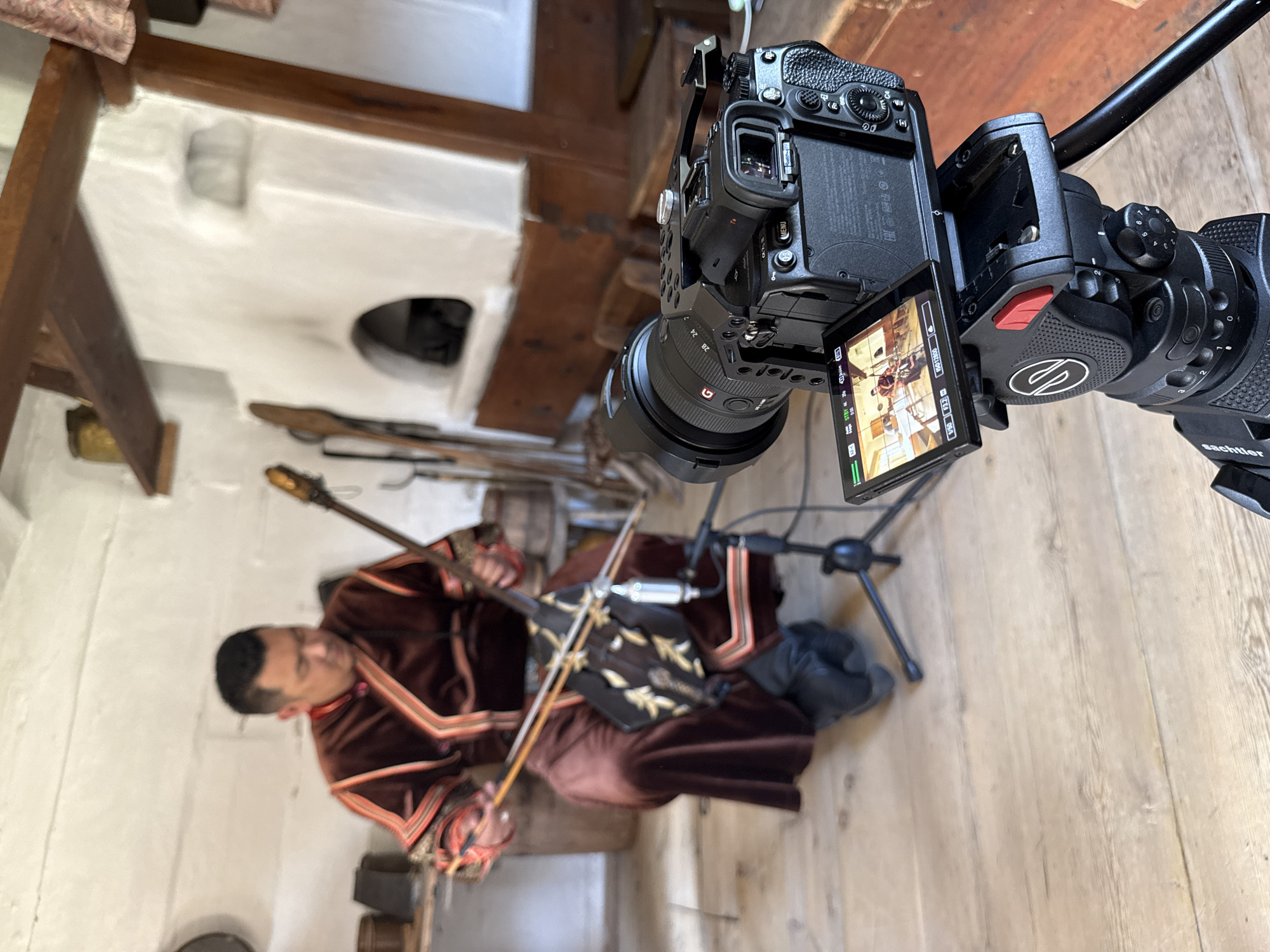 In a rustic room with whitewashed walls and wooden floorboards, a man dressed in traditional clothing, including a brown embroidered robe, is seated and playing a stringed instrument resembling a fiddle. He is focused on his playing, holding a bow in his right hand and the instrument in his left. In the foreground, a professional camera on a tripod is positioned to film the musician. The camera's screen shows a live feed of the performance, indicating a recording is in progress. The background reveals a traditional Russian stove and other rustic elements, suggesting a historical or cultural setting, possibly in the city of Shushenskoye, Russia, known for its historical significance and ethnographic museums.