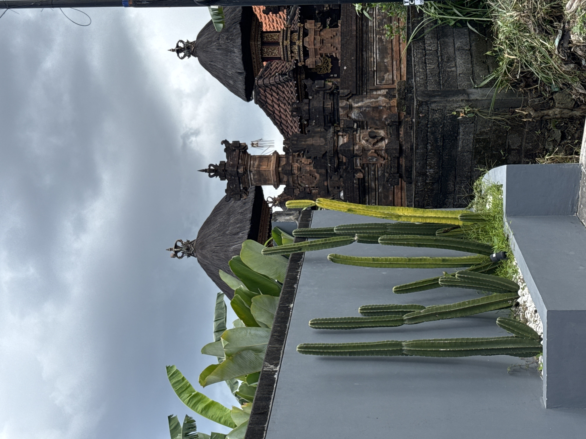 The image depicts a traditional Balinese architectural structure in North Kuta, Indonesia, set against a cloudy sky. The structure features tiered roofs with dark brown thatched tops and ornate details. A gray wall with cacti and green plants is in the foreground.