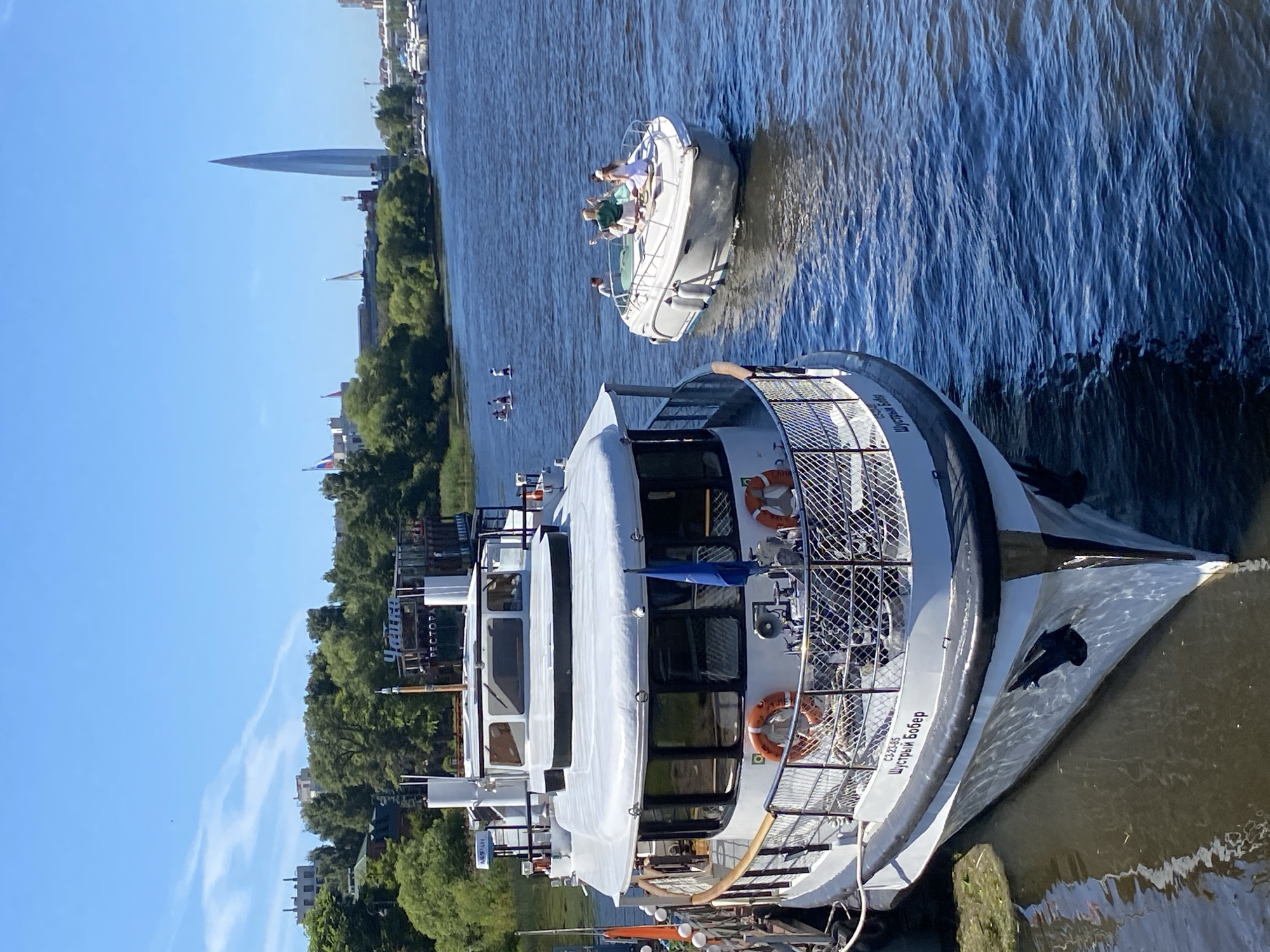 In Saint Petersburg, Russia, on a sunny day, the foreground features the bow of a large, white passenger ship docked at a pier. The ship has several windows and life preservers visible on its railing. The name "ШУСТРЫЙ БОБЁР" and the registration number "СЗ-23-85" are written on its hull. In the middle ground, a smaller white motorboat is on the water with four people on board. Three appear to be women, one wearing a green top and white skirt, another a white dress, and a third with a white hat. One person's gender is unclear. The water is blue and rippled. In the background, there is a lush green treeline along the bank of a river or canal. Beyond the trees, several buildings are visible, including a tall, slender, modern skyscraper with a pointed top, identifiable as the Lakhta Center, a prominent landmark in Saint Petersburg. A few other boats and structures are also discernible along the distant waterfront. The sky is clear and blue, with a few wispy white clouds. Several flags are flying from poles on the shore.