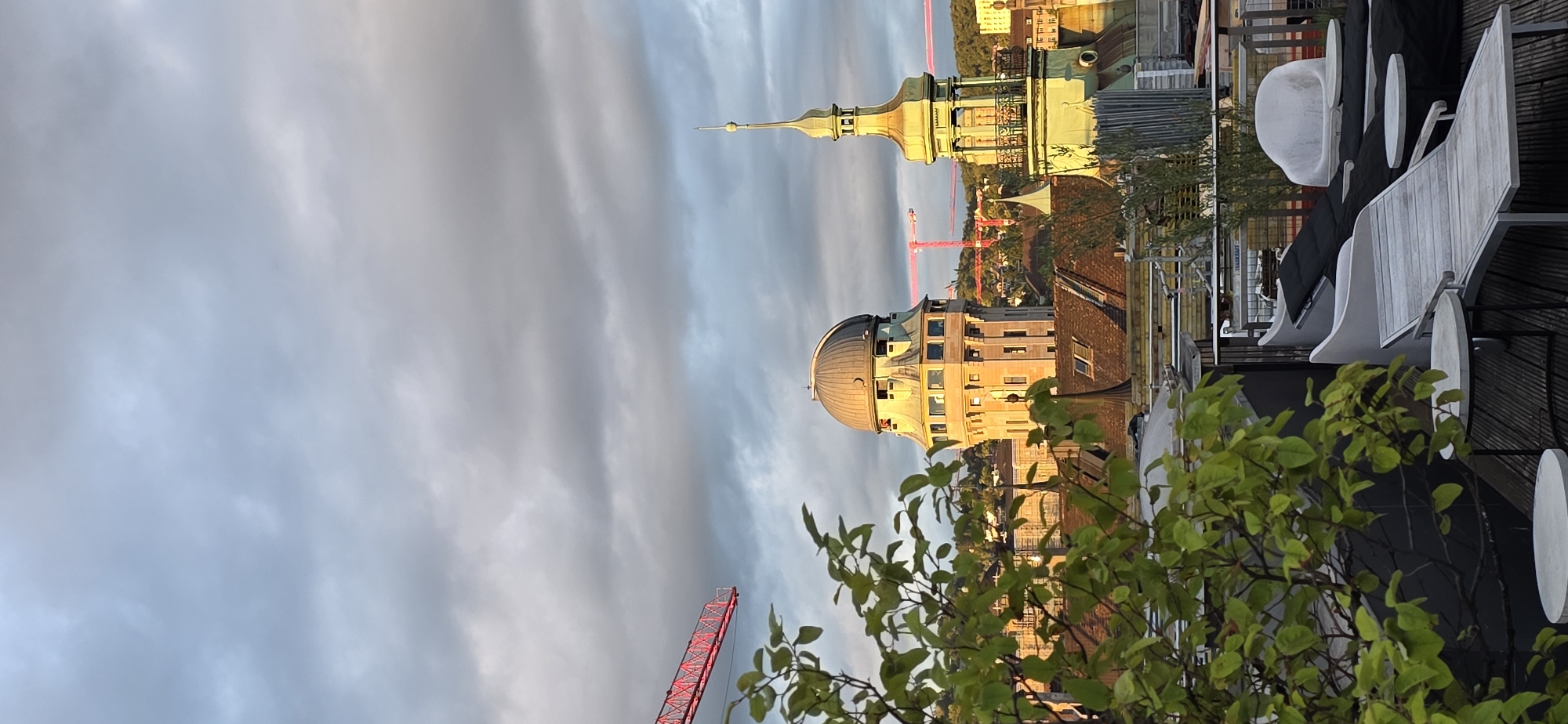 The image shows a rooftop view of a city, likely in Europe, with several notable buildings. In the center-left, a distinctive beige tower with a domed roof stands prominently, illuminated by warm sunlight. To its right, a green-spired tower with an ornate balcony rises. In the background, several red cranes are visible, suggesting ongoing construction. The foreground features a lush green tree with bright leaves on the left, and a seating area with modern loungers and small tables on the right. The sky is filled with dramatic, textured clouds, indicating either the early morning or late afternoon. The rooftops of other buildings, covered in dark tiles, are visible between the main structures. The overall scene depicts an urban landscape with architectural points of interest and a relaxed outdoor space.