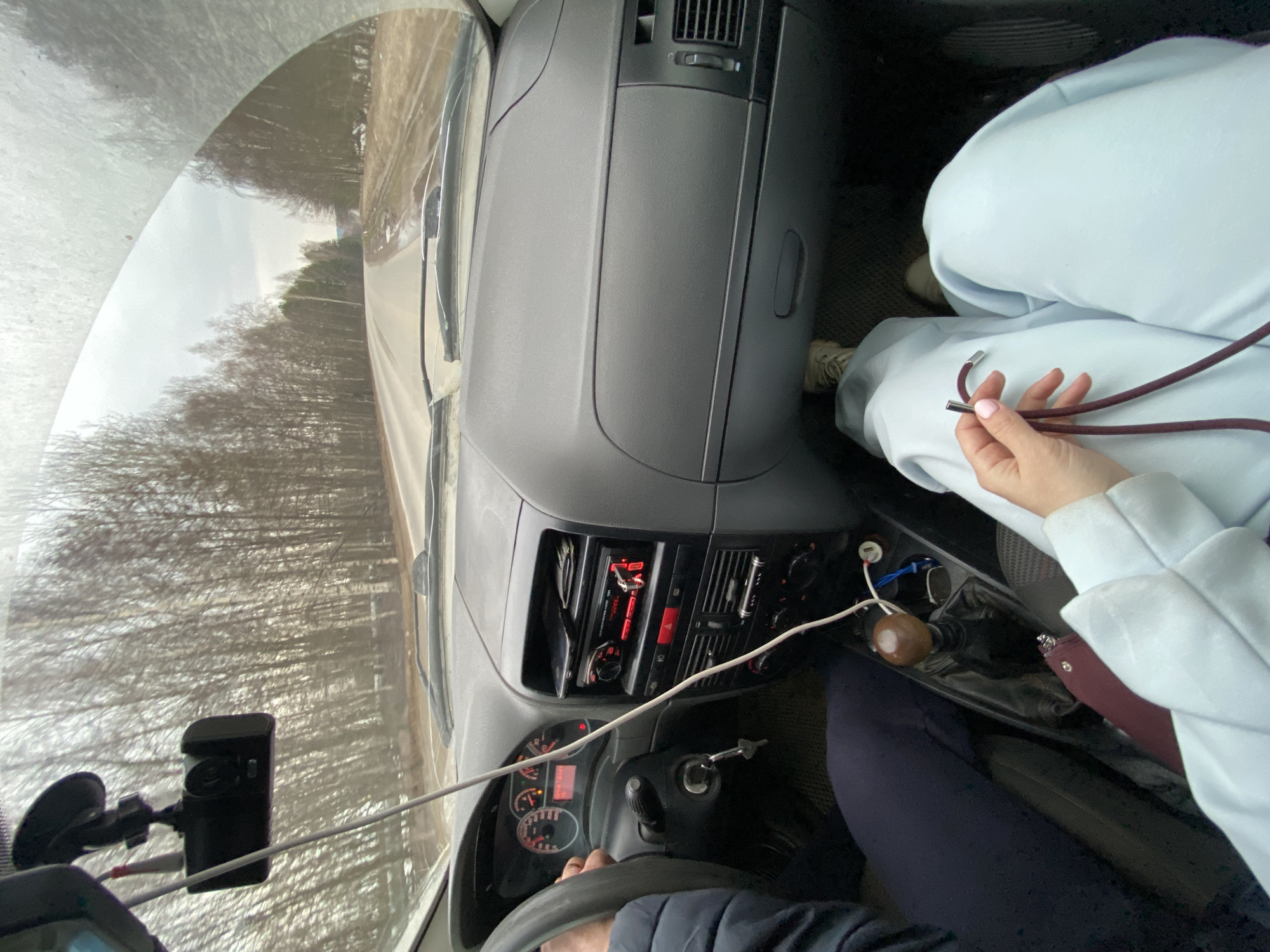 The image depicts the interior of a car, from the driver's perspective, suggesting motion based on the blurred background. The scene features two individuals: the driver and a passenger. The driver's hands are visible on the steering wheel. The passenger, wearing light blue clothing, is holding a dark red cord with metal ends. The dashboard and console are visible, with various controls and gauges. A dashcam is mounted on the windshield. Outside, trees line the road.
Based on the location provided, the scene takes place near Ol'khovskoye, Russia.