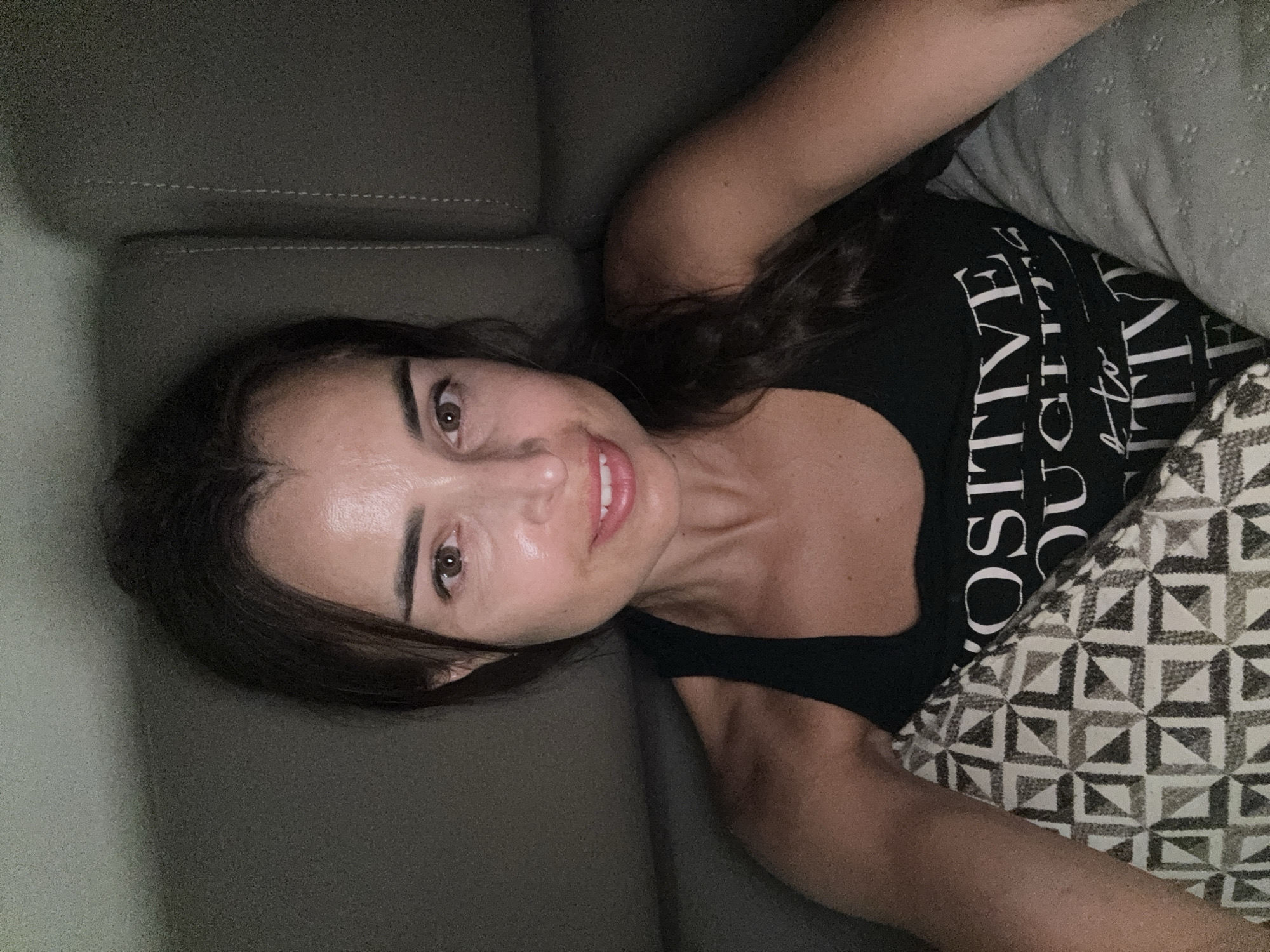 A woman is taking a selfie while sitting on a couch. She has brown hair pulled back and is wearing a black tank top with white text on it. She is holding a pillow with a geometric pattern. The background is a blurred couch.