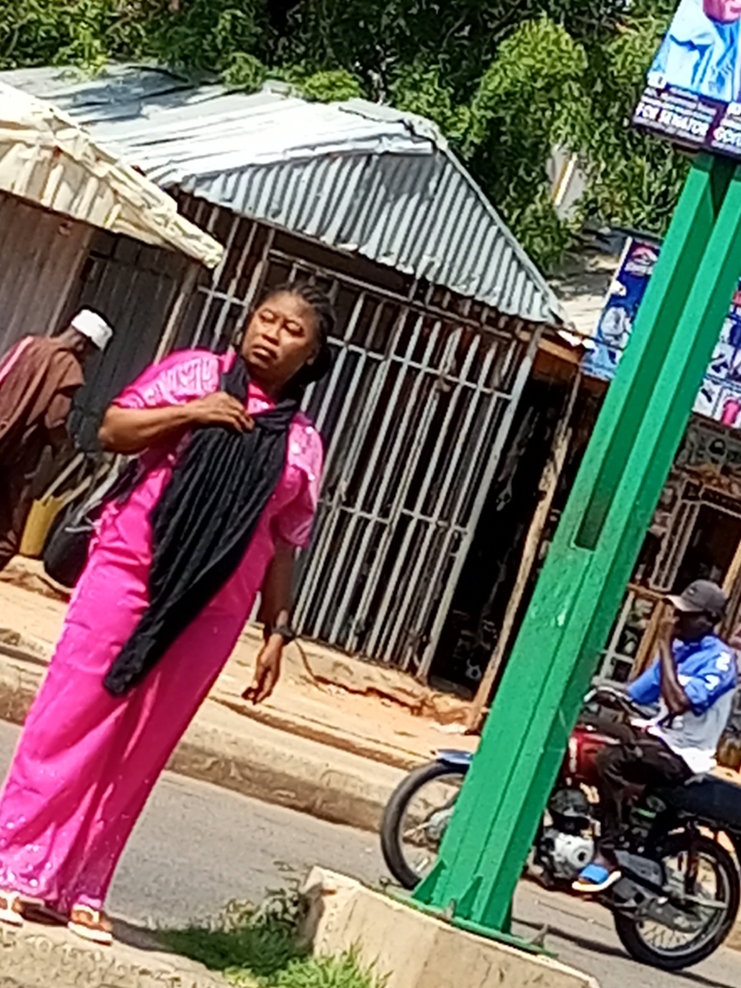 The image shows a street scene in Garko, Nigeria. In the foreground, a woman is walking, wearing a bright pink dress and a black scarf. She is carrying a black bag over her left shoulder. Behind her and to the left, another person is visible, wearing a brown robe and a white head covering. To the right of the woman, a green pole stands prominently, and behind it, a motorcycle is parked. A man in a blue shirt and dark pants is sitting on the motorcycle. Further in the background, buildings with corrugated metal roofs and barred windows are visible. There are also some election campaign posters attached to the green pole. The environment appears to be an outdoor, urban setting with some greenery.