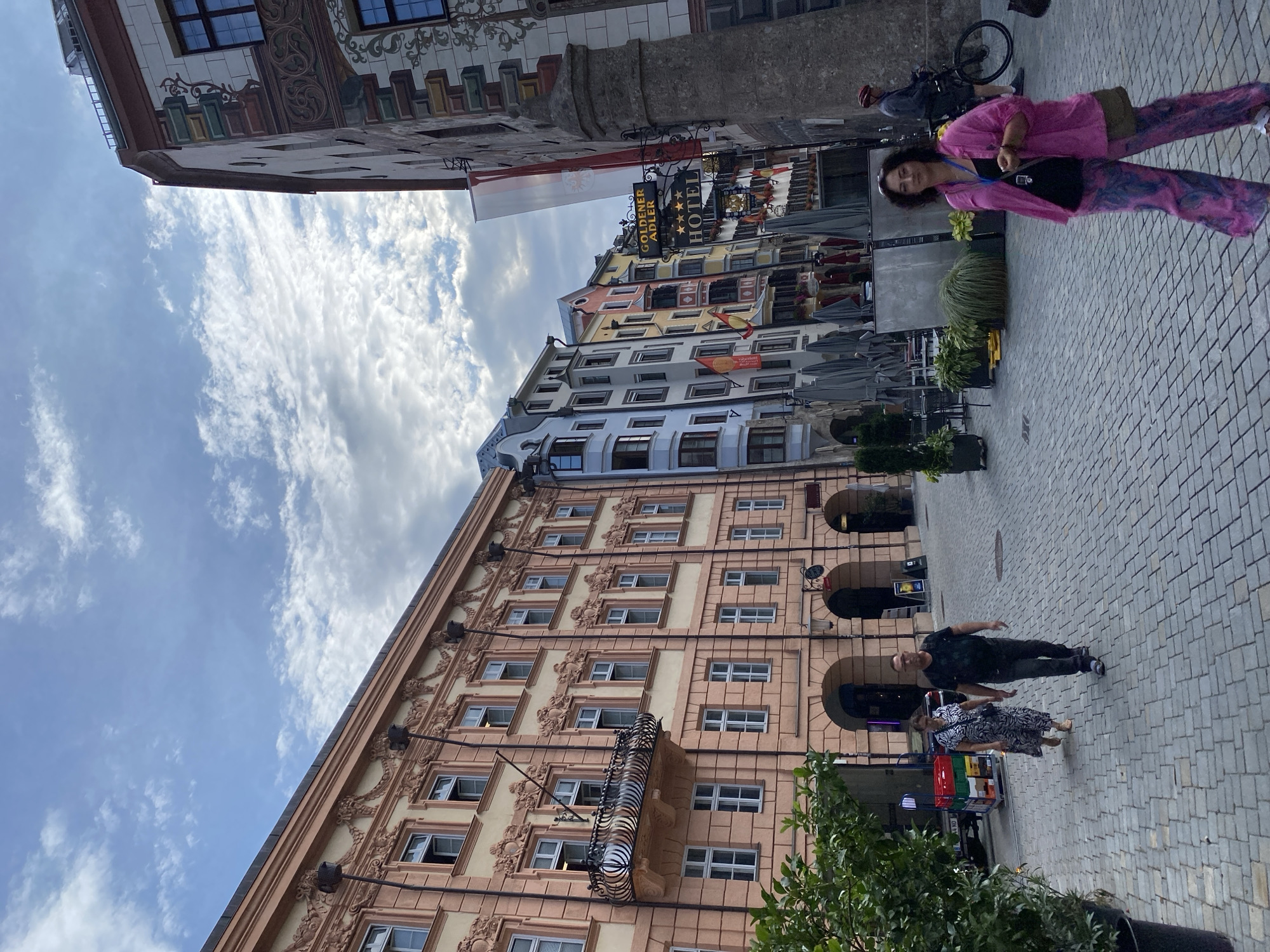 This is a street-level, eye-level, full-shot, outdoor, natural light photograph taken in Innsbruck, Austria, likely on the Golden Roof (Goldenes Dachl) street given the architecture and signage. The scene depicts a pedestrianized cobblestone street lined with colorful, historic buildings featuring ornate facades and balconies.
In the foreground, a woman with dark curly hair is walking towards the camera, wearing a bright pink open-front shirt over a dark top and patterned pink and blue pants. She has a crossbody bag and is holding a small object in her right hand. To the left, a couple walks away from the camera. The woman wears a patterned dress, and the man wears a dark t-shirt and jeans. Behind them, a delivery truck with a cart of crates is parked.
Further down the street, several other people are visible. One person on a bicycle is stopped on the right side of the frame. Outdoor seating with umbrellas is set up in front of some establishments. Signs for "GOLDENER ADLER HOTEL" with three stars are prominently displayed. The buildings showcase a mix of architectural styles, with pastel colors and intricate decorative elements. The sky above is partly cloudy.