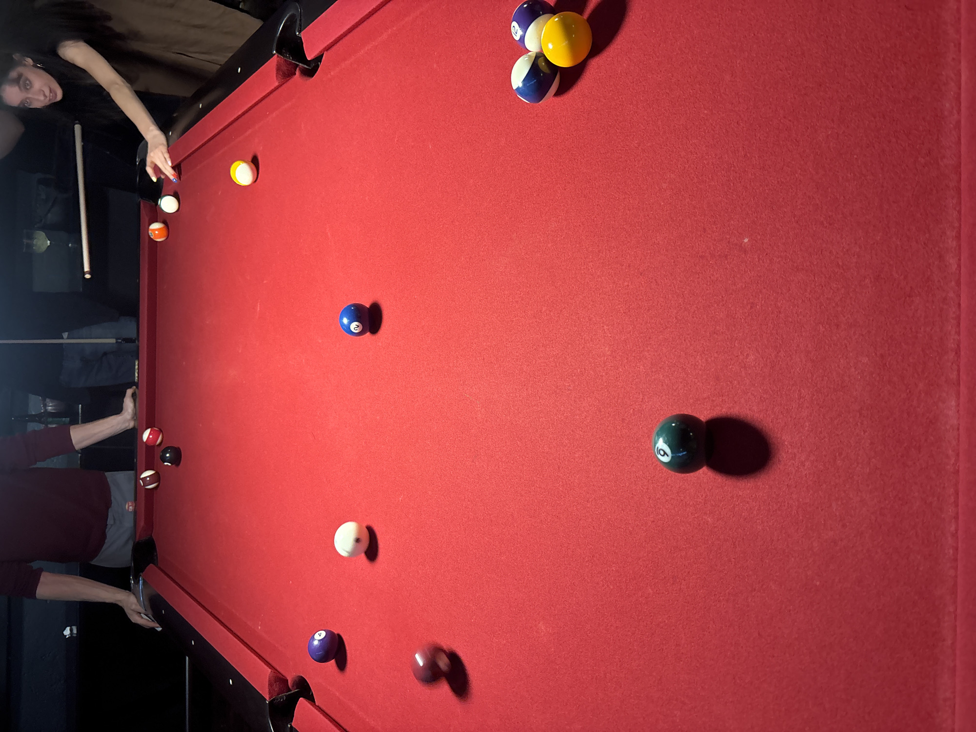 The image shows an overhead view of a billiards table, presumably indoors, with a red felt surface. Thirteen billiard balls are scattered on the table. A person is poised to strike the cue ball with a cue stick. Another person is on the left side of the table, also seemingly participating in the game. The lighting is ambient and the environment appears to be a recreational room. There is no information on the location in the city of Mexico City.