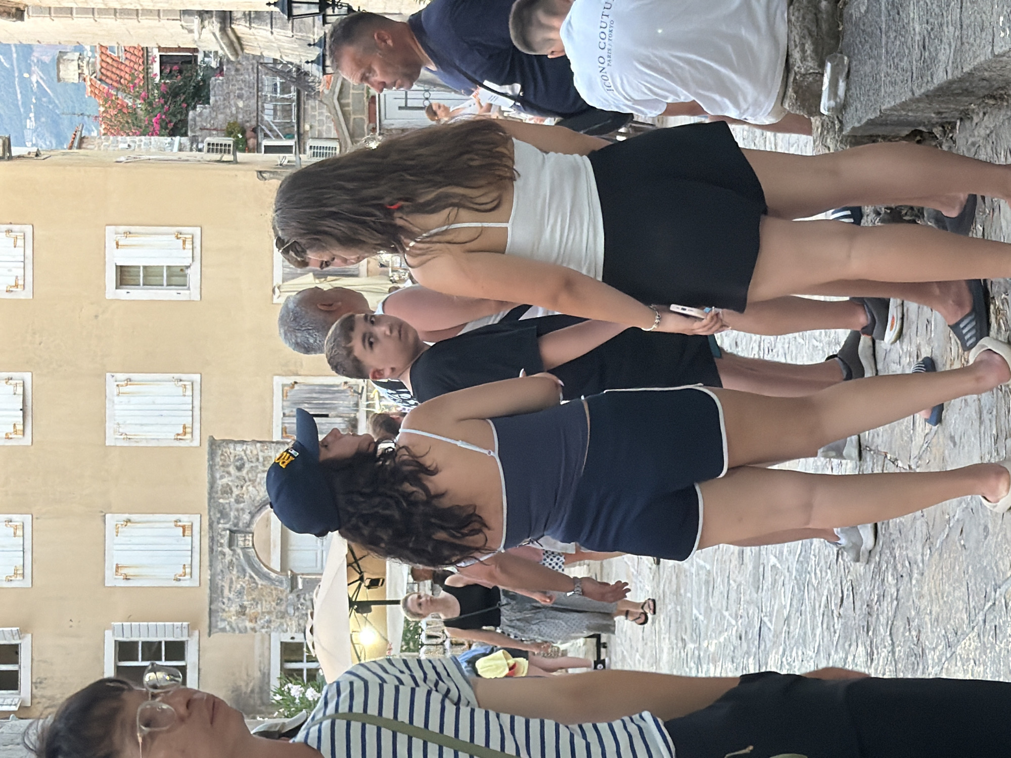 The image captures a bustling scene in the old town of Budva, Montenegro, with a focus on a group of people gathered on a cobblestone street. The architecture suggests a Mediterranean setting, with aged, light-colored buildings featuring shutters and balconies. In the foreground, a young person in a striped t-shirt is partially visible on the left, looking towards the center. Beside them, a young woman wearing a dark blue baseball cap and a matching tank top and shorts is engaged in conversation with a young boy. The boy, dressed in a black t-shirt, looks directly at the camera. To their right, another young woman in a white tank top and black shorts is holding a phone and appears to be interacting with her companions. Further in the background, an adult male in a blue t-shirt is visible, and another person in a white t-shirt with text is seated on a stone ledge. The overall impression is a casual gathering of people in a public space within a historic European town.