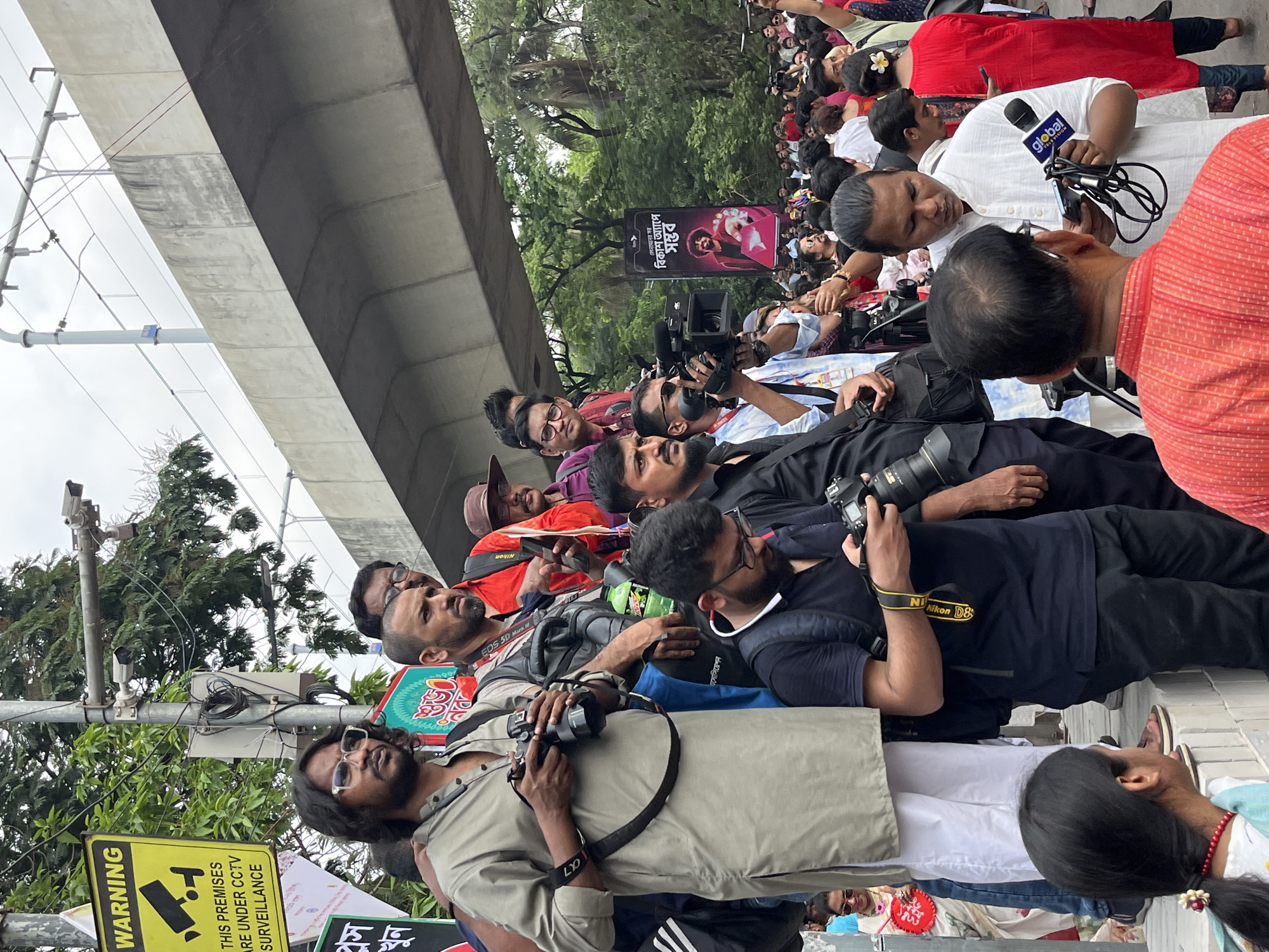 The image depicts a group of people, predominantly photographers and videographers, gathered on a sidewalk in what appears to be an urban environment. A concrete overpass looms overhead. The presence of a "Warning: These premises are under CCTV surveillance" sign indicates security measures in the area. A billboard in Bengali advertises " বিকাশ অ্যাপে সম্ভব " (Possible on bKash App), placing the scene in Bangladesh, specifically Dhaka due to the Bengali script. The people are focused on capturing images or videos, possibly of an event or subject out of frame. Some are using professional cameras and microphones. People wearing flower head ornaments lie down on the pavement. Overall, the scene suggests a media event or public gathering in a city street.