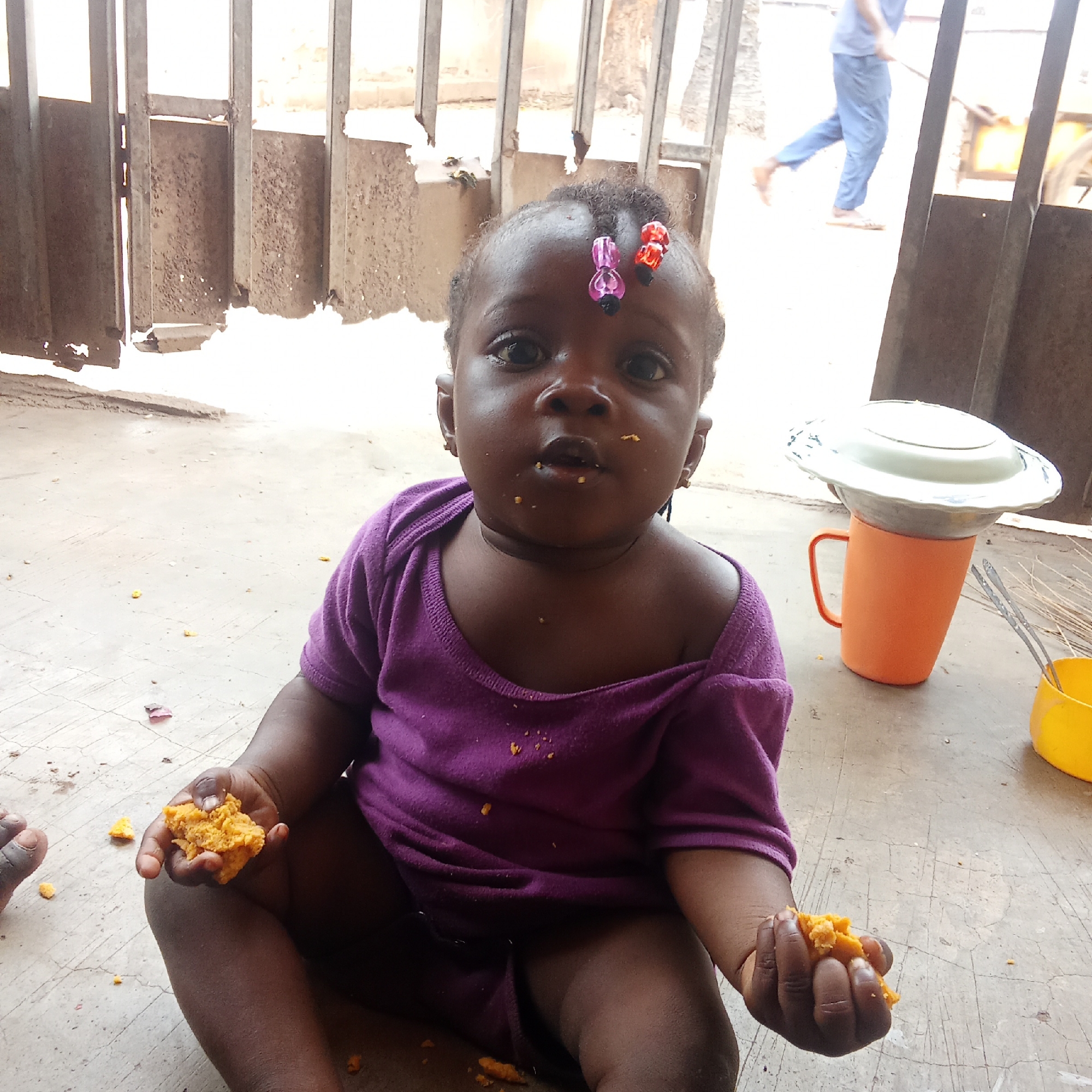 Akwanga, Nigeria. A dark-skinned baby sits on a concrete floor wearing a purple shirt, eating pieces of orange food. The baby has hair clips in their hair and is surrounded by scattered crumbs. Behind the baby is a metal gate and a blurry figure in blue pants. To the right of the baby are an orange mug and a yellow cup.