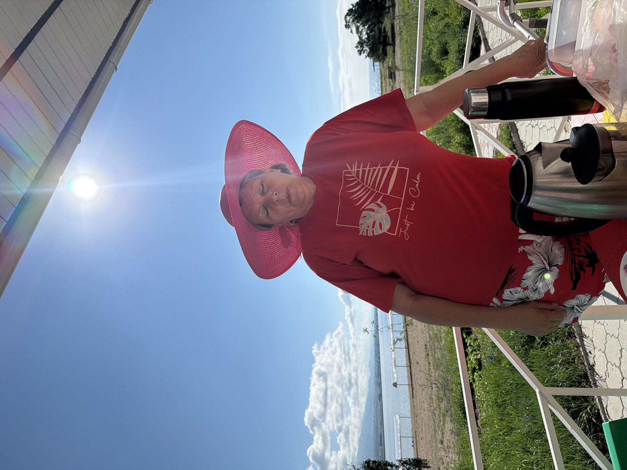 A woman in a red t-shirt and red floral shorts stands on a balcony overlooking a body of water. She is wearing a large, floppy pink hat and has her eyes closed. In front of her, on a table, are a silver electric kettle, a black thermos, and a clear plastic container. The sky is bright blue with a few scattered clouds, and the sun is shining. In the background, there is a beach, trees, and distant hills. The woman appears to be enjoying a peaceful moment outdoors.