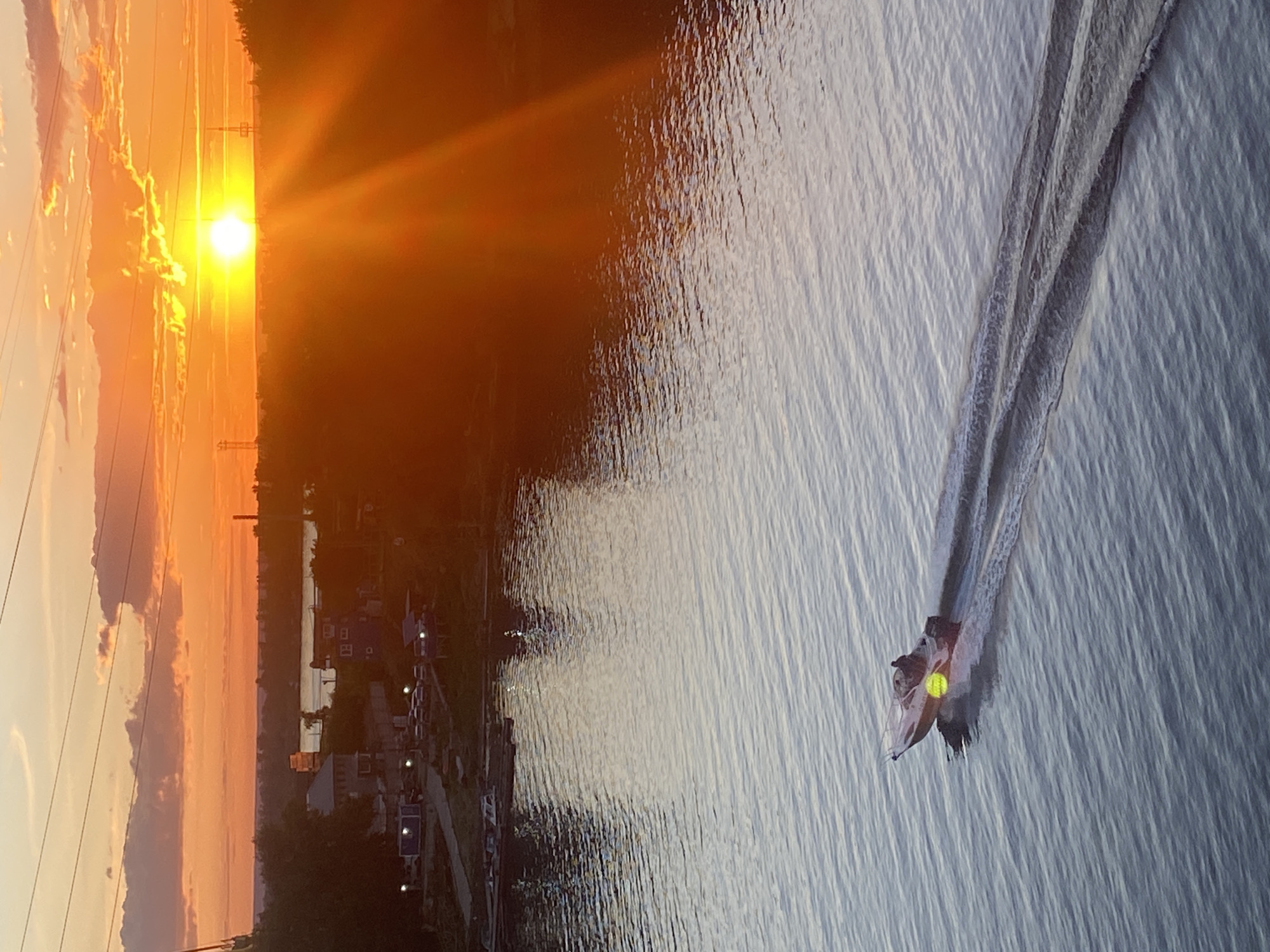 A vertical, high-angle shot captures a serene sunset over a body of water. The sun, a brilliant orb of golden light, is low on the horizon, casting a warm, orange glow across the sky and reflecting on the rippling surface of the water. Wispy clouds are painted in shades of orange and pink, adding depth to the dramatic sky.
Along the shoreline, silhouetted against the vibrant sunset, is a small town with various buildings and structures. Barely visible, a few lights are beginning to twinkle from the houses. A line of utility poles with power lines extends across the left side of the frame, leading towards the horizon.
In the foreground, a small boat is cutting through the water, leaving a white wake in its path. Two individuals are visible in the boat, their forms indistinct in the dimming light. The water itself is a mosaic of light and shadow, with the sunset's reflection creating streaks of warm color interspersed with the cool tones of the water. The overall mood is peaceful and tranquil, evoking the end of a day in a quiet, possibly rural, location. The scene suggests a late afternoon or early evening, with clear weather.