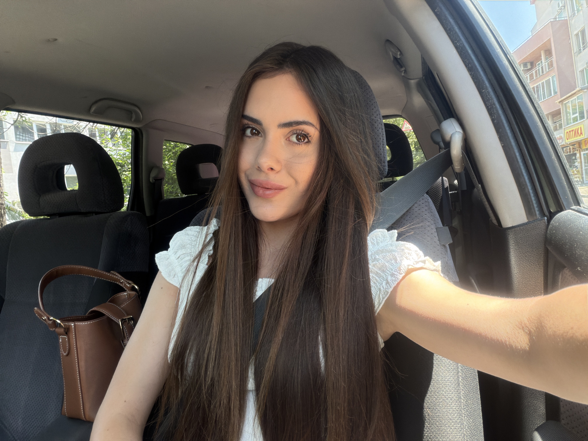 A young woman with long, dark brown hair and brown eyes is captured in a selfie taken inside a car. She is looking directly at the camera with a subtle, pleasant smile. She wears a white top with ruffled sleeves, and a seatbelt crosses her chest. The car's interior, featuring dark fabric seats and headrests, is visible behind her and to her left. A brown, structured handbag with white stitching is placed on the seat beside her.
Through the car windows, the exterior environment is visible, indicating a sunny daytime scene with clear blue skies. Lush green trees and multi-story buildings are in the background. A prominent detail is a storefront sign on one of the buildings, displaying the Cyrillic word "ОПТИКА" (OPTICA), which translates to 'Optics' or 'Optician's'. This text, along with the given context, suggests the location is in Burgas, Bulgaria. The strong sunlight casts shadows, confirming the bright weather conditions. The woman's relaxed posture and soft smile suggest a calm and positive mood during the event.