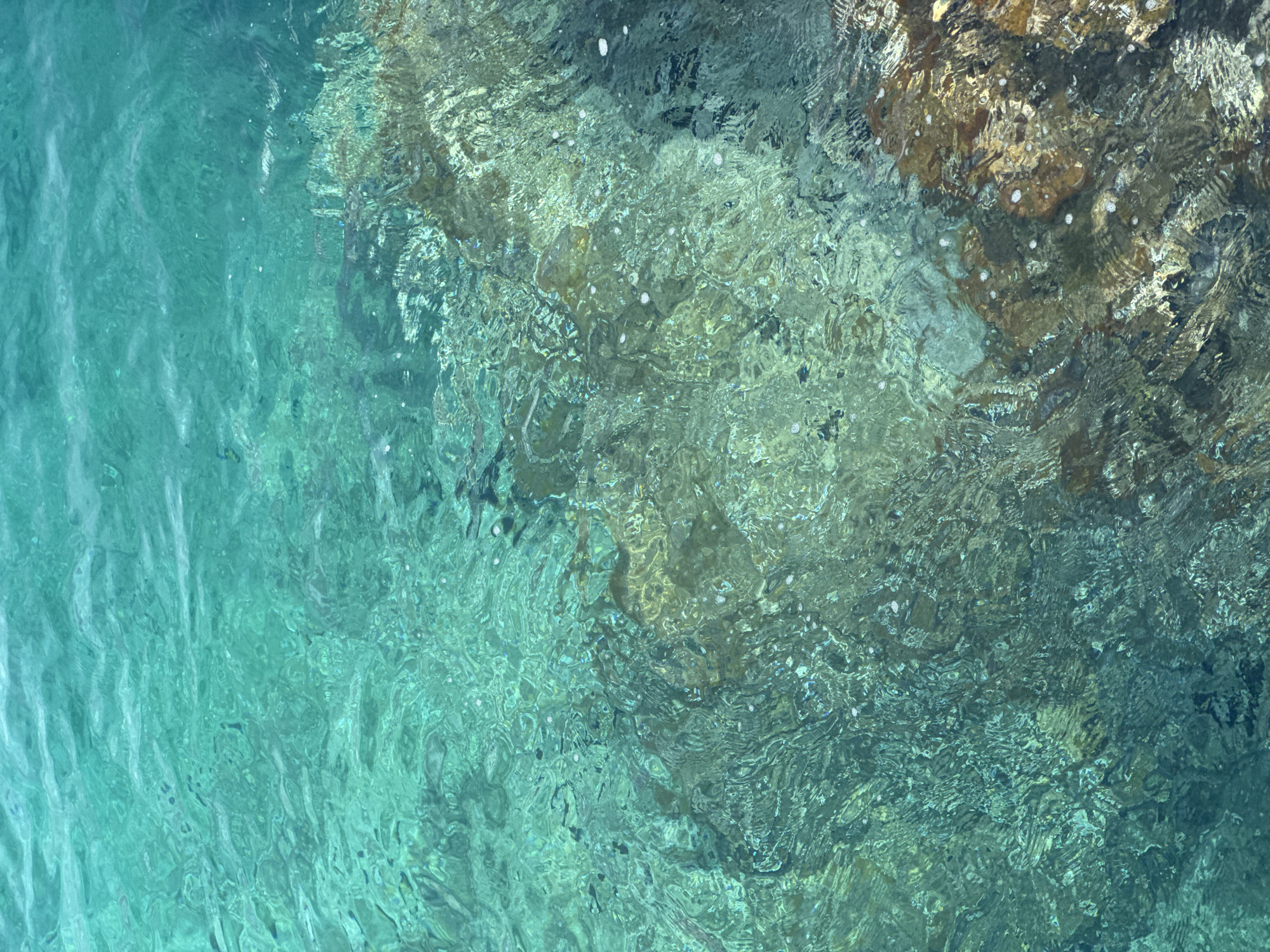 This media file is an abstract, high-angle, full shot of shallow, clear, turquoise water flowing over rocks. The water is in constant motion, creating ripples and distortions that obscure the details of the underwater rocks and seabed. The light refracts through the water, creating a shimmering effect with bright highlights and darker shadows. Small white bubbles are scattered throughout the water's surface, particularly in areas where the water is more agitated. The rocks visible beneath the surface are varied in color, ranging from dark grey to brown and yellow, with some appearing to be covered in algae. The scene is illuminated by natural light, suggesting it is daytime. There is no visible text in the image. The setting appears to be a natural body of water, likely a coastline or riverbed, given the rocky substrate. The overall impression is one of natural beauty and the dynamic interplay of light and water.