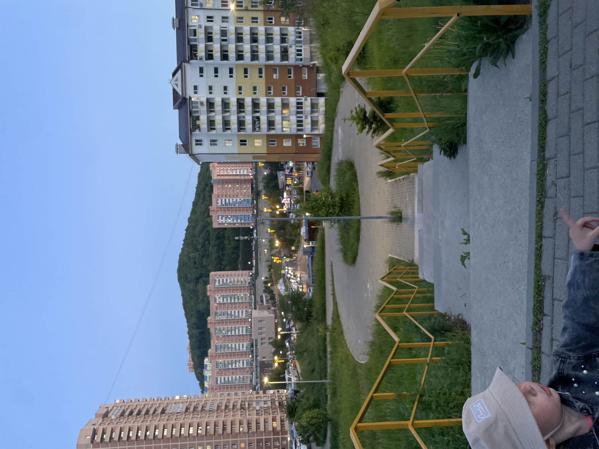 The image shows an urban landscape in Vladivostok, Russia. In the foreground, a set of concrete stairs with yellow metal railings leads down towards a paved pathway. A child wearing a beige bucket hat and a denim jacket is visible in the lower right corner, partly obscured.
Beyond the pathway, several multi-story residential buildings of varying colors (beige, brown, white) line the street, with lights visible in some windows. Streetlights illuminate the road, indicating it is evening or dusk. A densely wooded hill forms the backdrop behind the buildings. The sky is a pale blue, suggesting twilight. The overall scene captures a residential area with pedestrian access via the stairs, with natural and built environments juxtaposed.