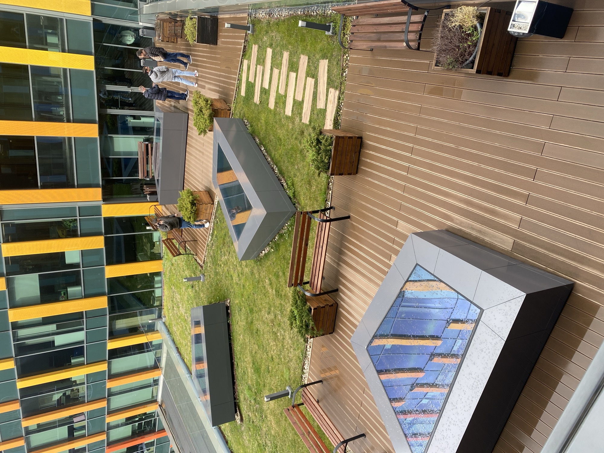 The image shows an outdoor recreation area in a modern building complex. There is a combination of wooden decking, grassy areas, and rectangular planters. Benches are provided for seating. Several people are present, some standing and some sitting on benches. The architecture is modern with a glass and metal building facade visible. A large square structure with a glass top, possibly a skylight, dominates the foreground. The image suggests an outdoor space designed for relaxation and socializing within a modern urban setting. Given the architecture and landscape elements, it is plausible that this is in the city of Sofia, Bulgaria.