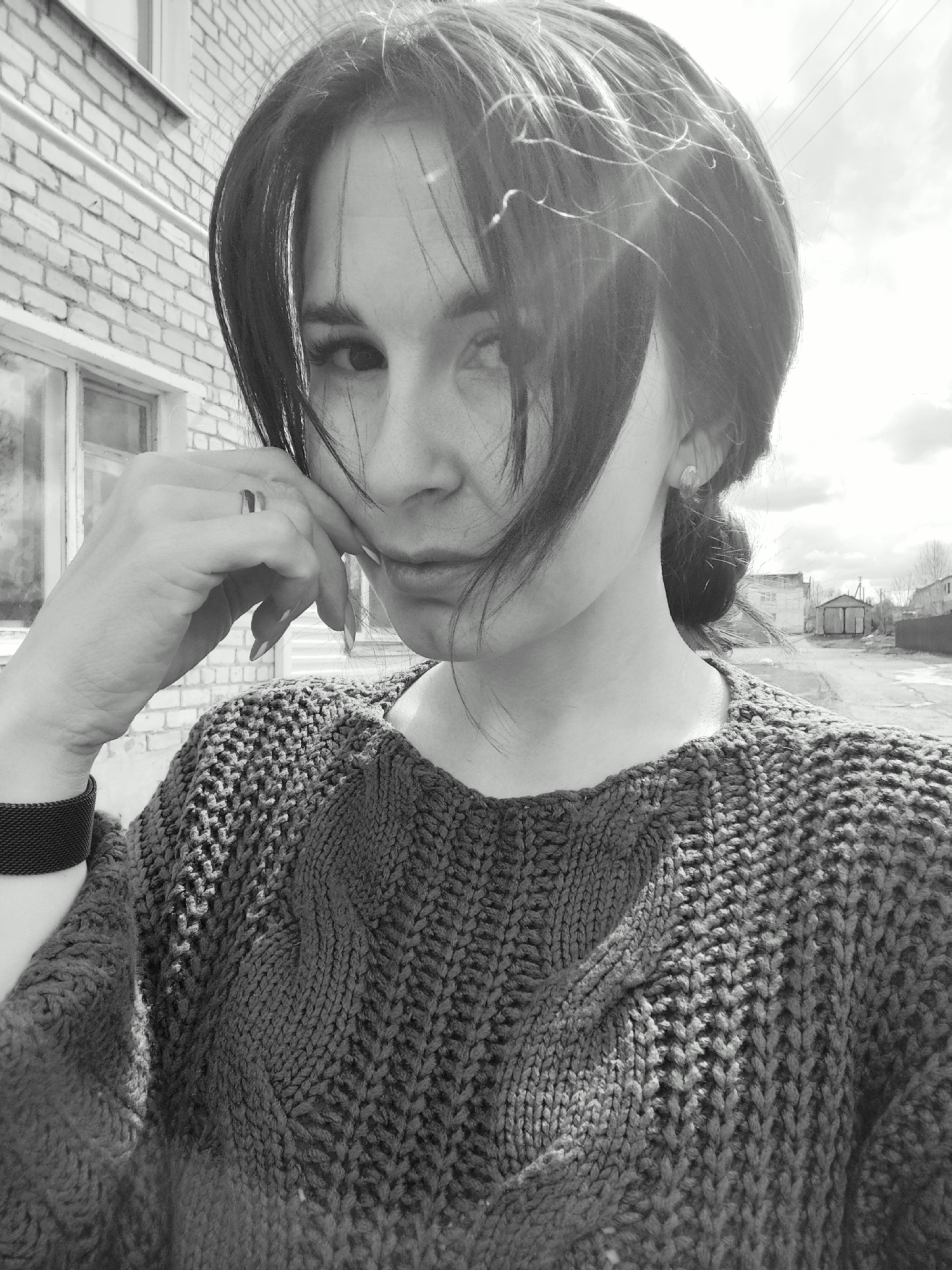 A black and white, eye-level, medium indoor/outdoor shot shows a woman with dark hair in front of a brick building. The woman’s face is partially obscured by her hair and her hand which she is holding to her cheek. She wears a dark, heavy knit sweater and a dark bracelet. The brick building is behind her on her left. The background on her right shows a street with buildings. The buildings in the background are identified as being in the city of Фоки, Russia.