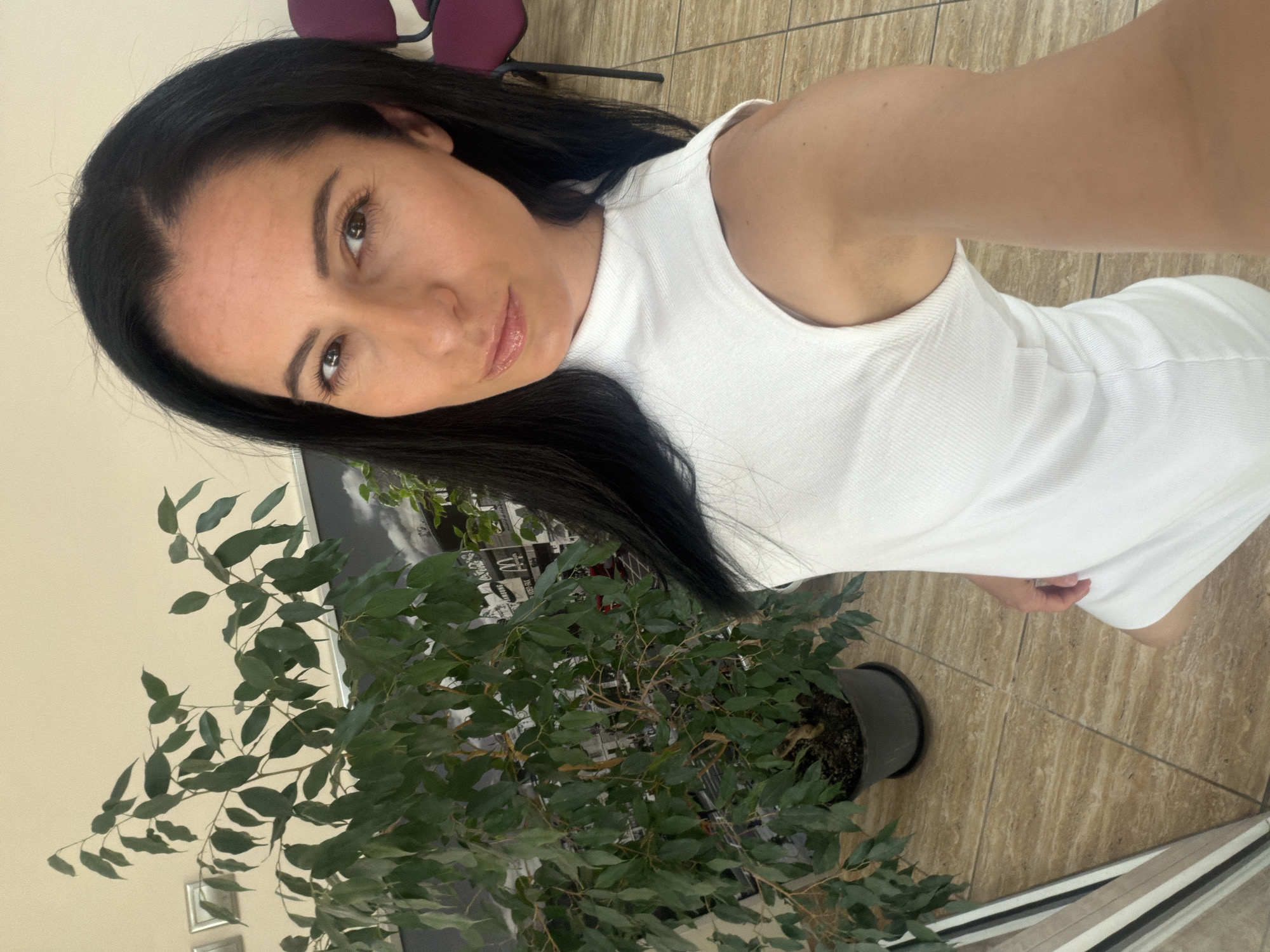 A woman with dark hair and brown eyes is posing for a selfie. She is wearing a white sleeveless dress. A large potted plant with green leaves is to her left. The background appears to be an interior space with tiled flooring. A partial view of a purple chair is visible in the upper left corner. The location is Pleven, Bulgaria.