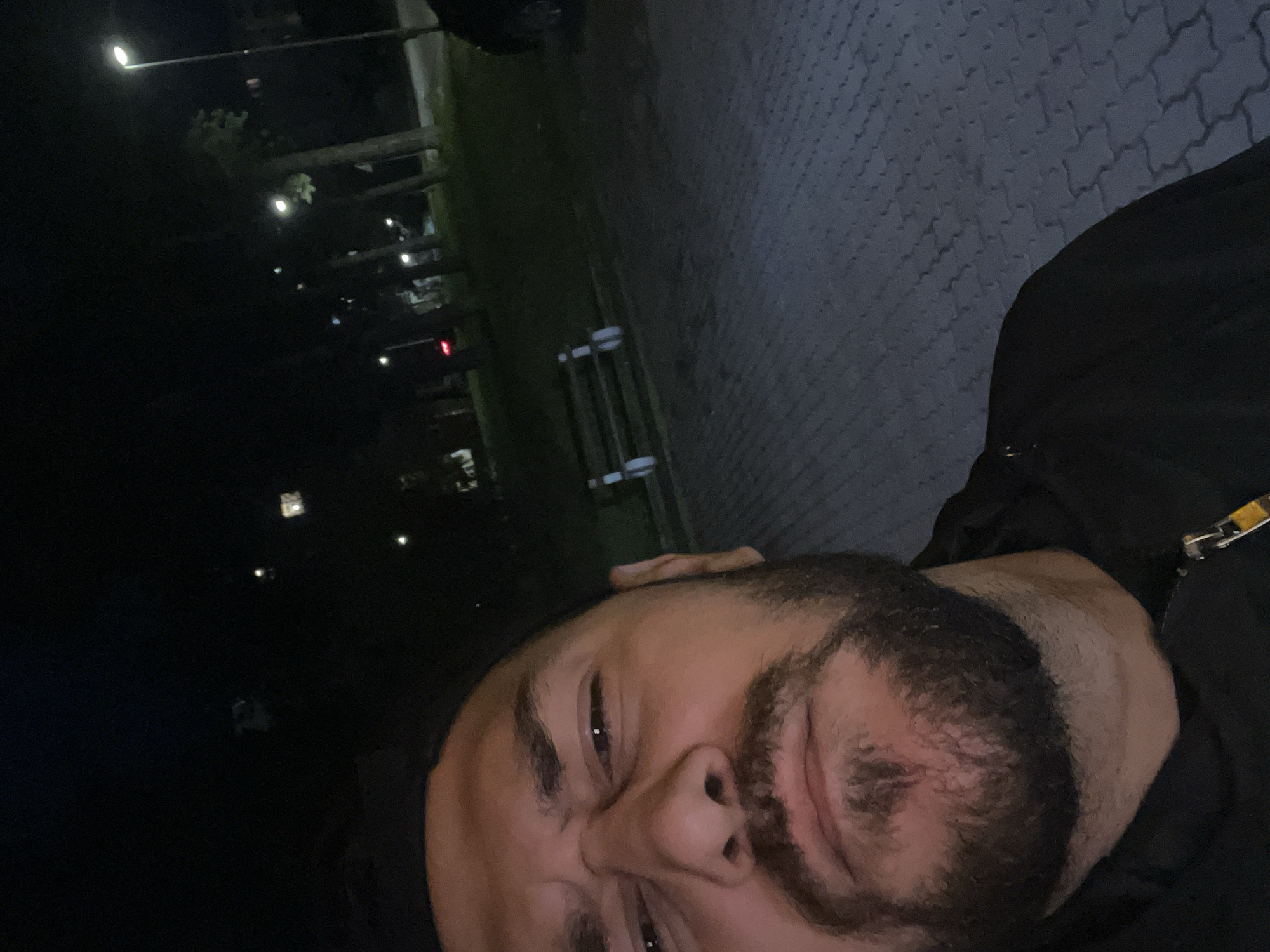 A selfie is taken at night in Veliko Tarnovo, Bulgaria. The individual has a short beard and is wearing a dark jacket. Street lights are visible in the background. The ground is paved with interlocking blocks.