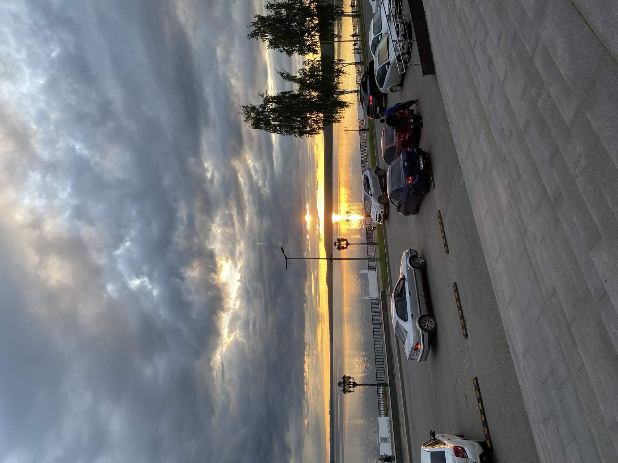 The image depicts a sunset scene in Votkinsk, Russia, alongside a body of water, likely the Votkinsk Reservoir. The sky is filled with dark, dramatic clouds tinged with orange from the setting sun. The sun's reflection is visible on the water's surface.
A row of trees lines part of the waterfront. Along the roadside, several cars are parked. Period streetlamps stand along the edge of the water. A gray concrete wall runs on the right side of the frame.