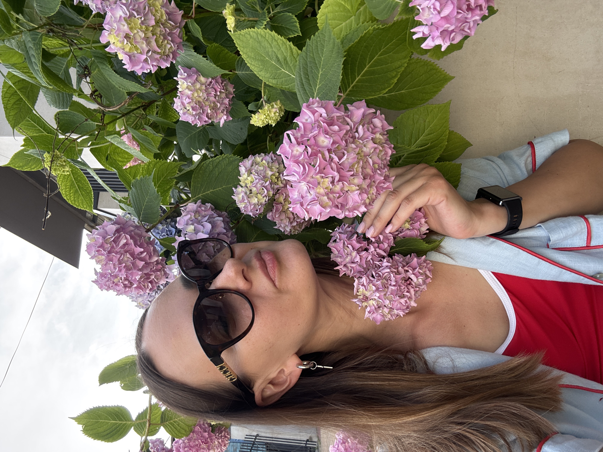 A woman, appearing to be in her late 20s or early 30s, with shoulder-length brown hair, is posing for a selfie in front of a wall and a dense bush of pink hydrangeas. She is wearing large, dark sunglasses with the "GUCCI" logo visible on the side, a silver earring in the shape of a key, a red tank top, and a light blue jacket with red trim. She is touching a cluster of hydrangeas with her right hand. The background shows a beige wall, a metal railing, and a building with a dark roof. The scene appears to be outdoors, likely in Sochi, Russia, given the floral environment and architecture. The sky is overcast.