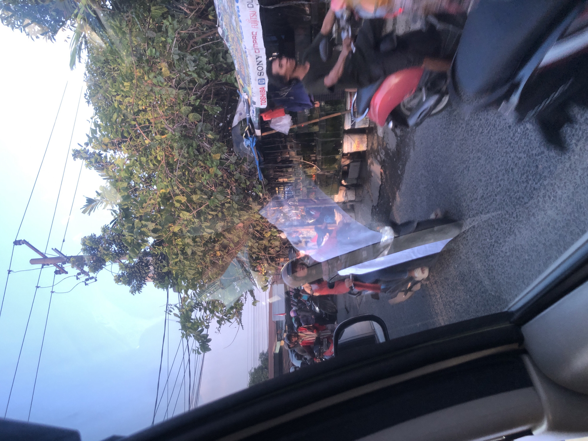 This image captures a street scene in Medan, Indonesia, likely during daylight hours, with a view from inside a vehicle. The foreground shows the interior of a car, including the side mirror and window frame, with reflections on the glass. The road ahead is populated with several motorcycles and their riders, suggesting traffic or a busy street.
A man on a red scooter is prominently featured, wearing a dark t-shirt and black pants. He is looking to his right. Behind him, other individuals on motorcycles are visible, some wearing helmets. One woman on a scooter wears a red jacket and a helmet, and her face is partially obscured by a transparent windshield. Another motorcyclist in the middle ground wears a striped shirt.
The environment includes a roadside stall with a blue tarp and a white banner displaying logos such as "TOSHIBA," "SONY," and "FUJITSU." Several buckets are placed on the ground near the stall. Overhanging trees with lush green foliage line the street, and power lines are visible against a bright sky. The road surface appears wet in places, indicating recent rain or a damp condition. The overall atmosphere suggests a typical urban setting in Medan, characterized by motorcycle traffic and street vendors.
