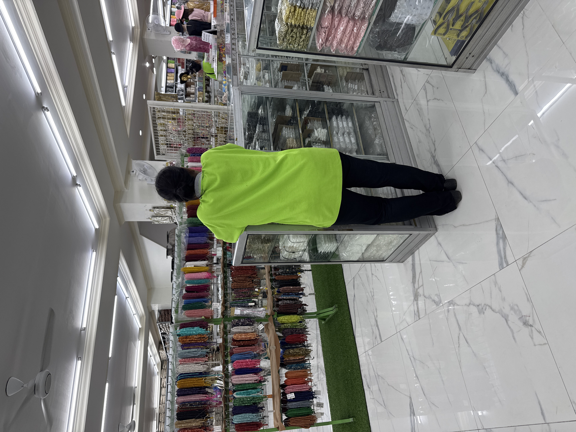Inside a brightly lit retail store in Kuta, Indonesia, a person with dark hair stands facing a glass display case. They wear a neon green short-sleeved shirt and dark pants. To their left, shelves display multiple strands of colorful beads. Above and to the right are glass display cases filled with items, presumably for sale. The floor is white tile with grey marbling. Fluorescent tube lights illuminate the ceiling, and several ceiling fans are visible.