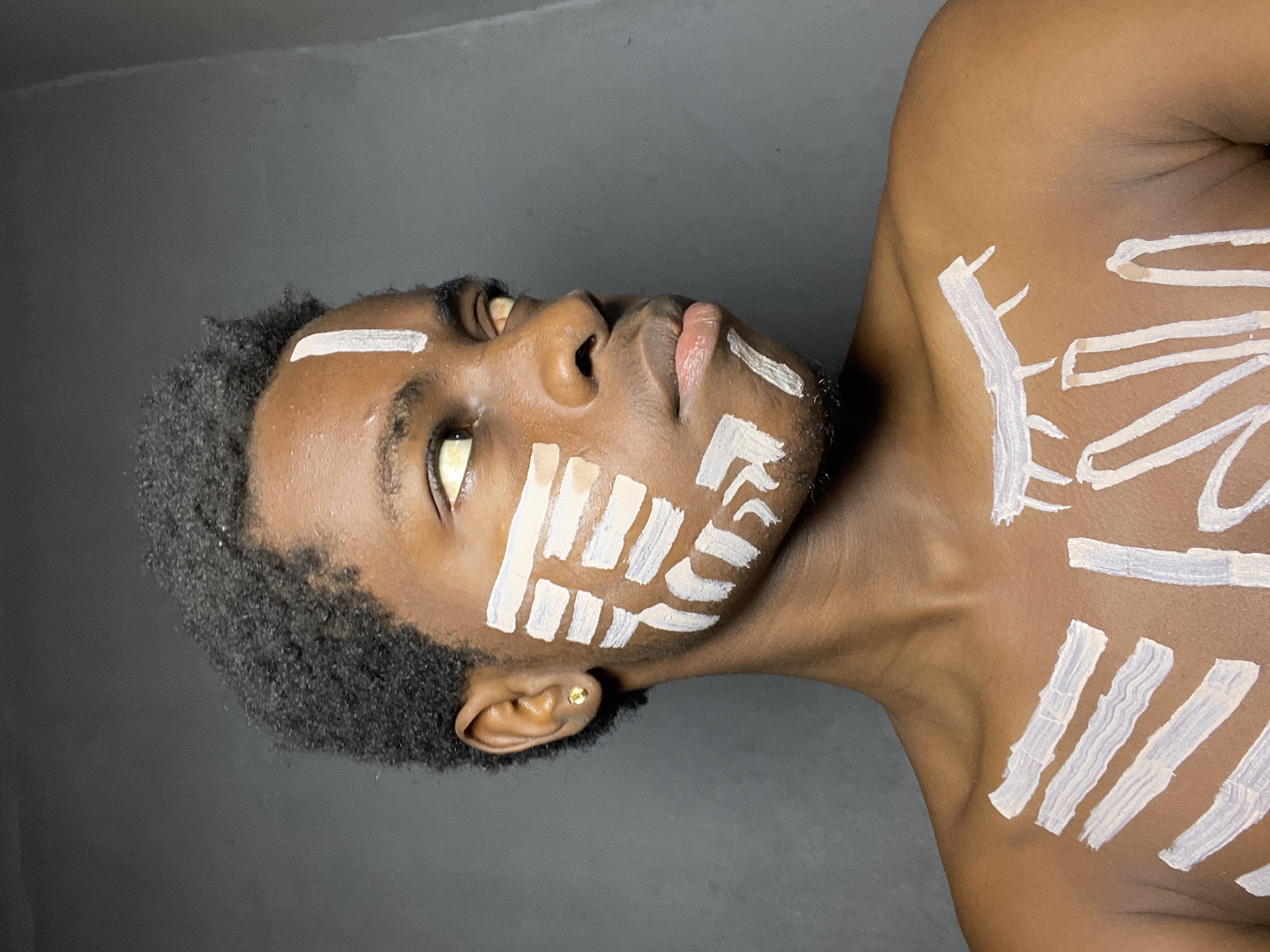 A low-angle portrait shows a dark-skinned, bare-chested man with close-cropped, dark, natural hair and white markings in horizontal strokes on his face and chest. He is looking upward. He has a gold stud earring in his left ear. The background is a dark grey or black color. The location is unknown but presumed to be in Nkpor, Nigeria.