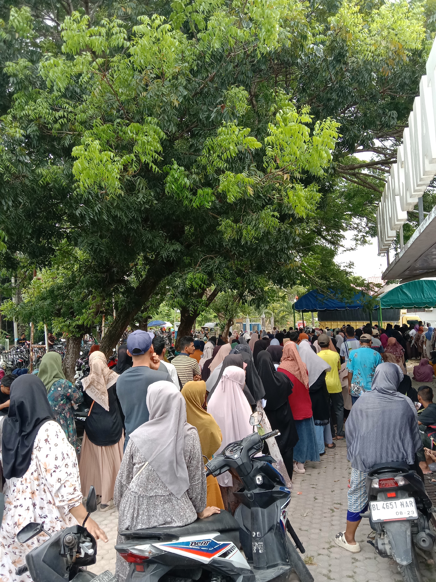 The image depicts a dense crowd of people, predominantly women wearing headscarves (hijabs), standing in a line in Lhokseumawe, Indonesia. The crowd extends from left to right, with some people appearing to be seated on motorcycles in the foreground. The license plate on one motorcycle is visible, reading "BL 4651 NAR 08-29." A large tree with thick foliage provides shade over much of the area where the crowd is gathered. In the background, there are glimpses of tents and other structures, suggesting an event or distribution of some kind. The ground is paved with interlocking concrete pavers.