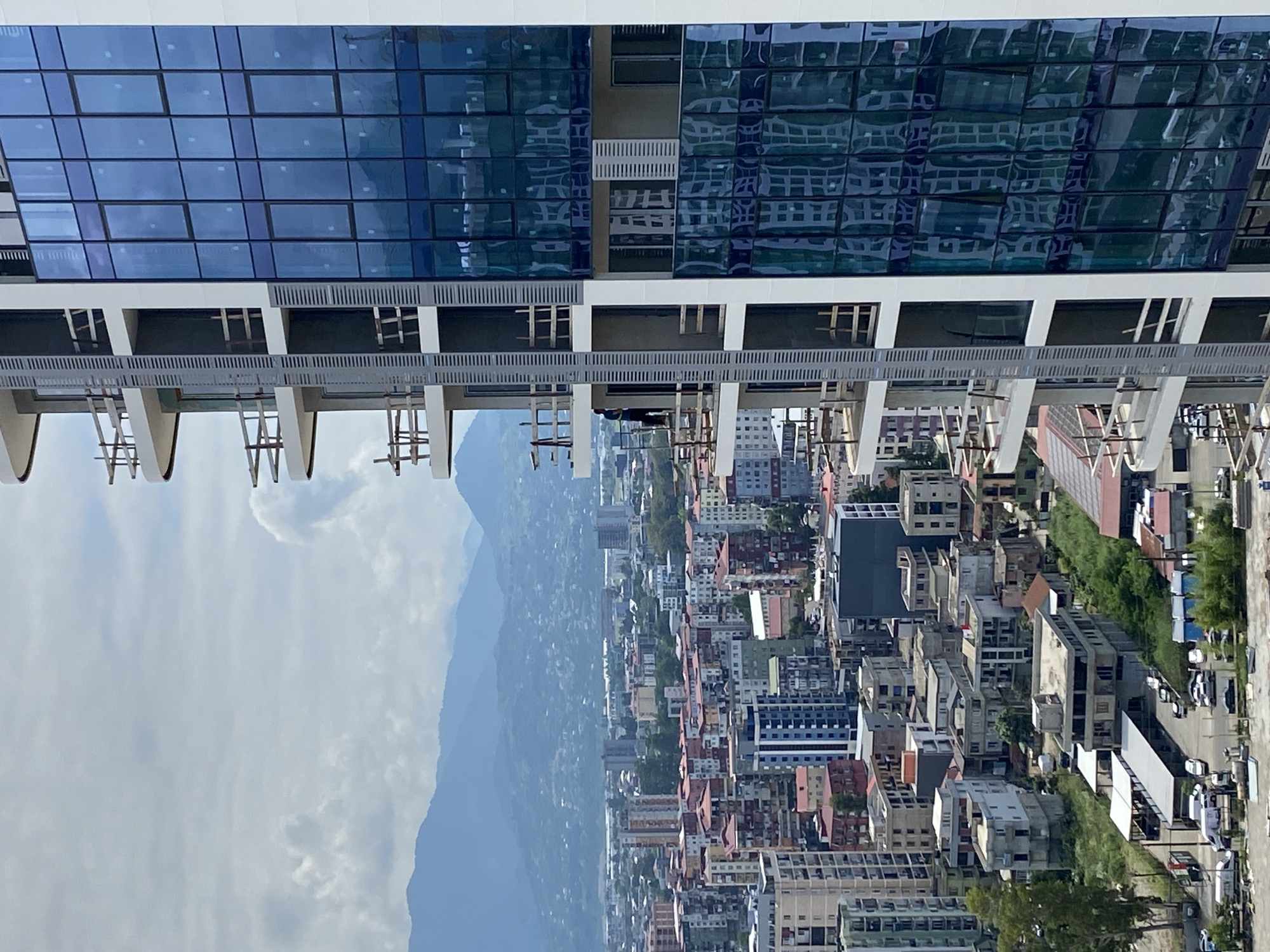 This image captures a section of the Batumi coastline, focusing on a modern skyscraper under construction alongside older, multi-story buildings. The skyscraper, on the right side of the frame, features a facade of blue glass panels, reflecting the sky and surrounding cityscape. Balconies with wooden scaffolding are visible on the left side of the skyscraper, indicating ongoing construction work. A lone figure, appearing to be a construction worker, is visible on one of the balconies.
The older buildings in the mid-ground are a mix of architectural styles and colors, with some exhibiting red tiled roofs. The urban landscape extends towards a range of misty, blue mountains in the background, suggesting the city's location near the Caucasus Mountains. The sky is overcast with visible clouds. In the foreground and to the left, a lower-rise area with parking lots and smaller structures is present, with some vehicles and shipping containers visible. The overall scene depicts a transitional urban environment, blending new development with established neighborhoods against a natural backdrop.