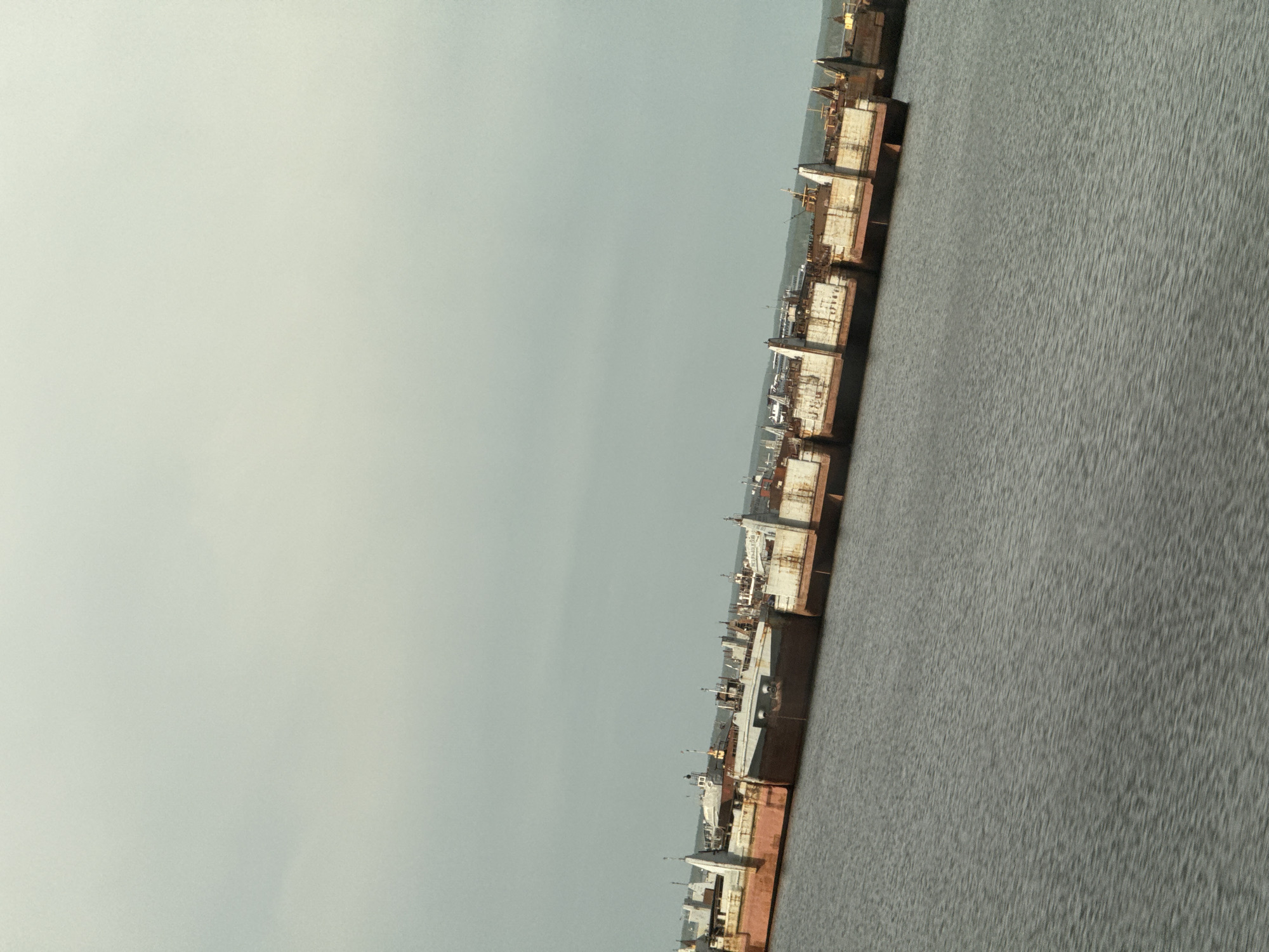 The photograph depicts a line of rusting ships anchored in a harbor. The sky is overcast. Ripples are visible on the water. The city is likely located in the State of Unknown and City of Unknown.
