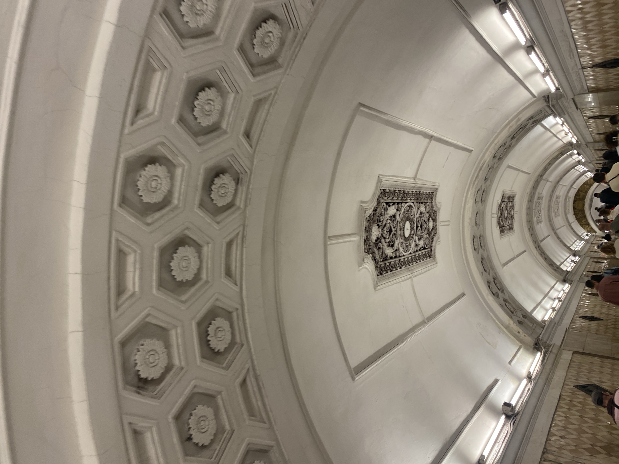 The image shows the ceiling of a Moscow Metro station, characterized by its grand architectural style. The ceiling features a series of hexagonal coffers, each containing a decorative floral motif. In the center of the image, a larger, ornate decorative panel is visible within a recessed rectangular frame. The walls of the station are lined with decorative tiles and illuminated by fluorescent lighting. Several people are walking along the station's corridor, suggesting it is in use by commuters. The overall impression is one of opulence and historical significance, typical of many stations in the Moscow Metro system.