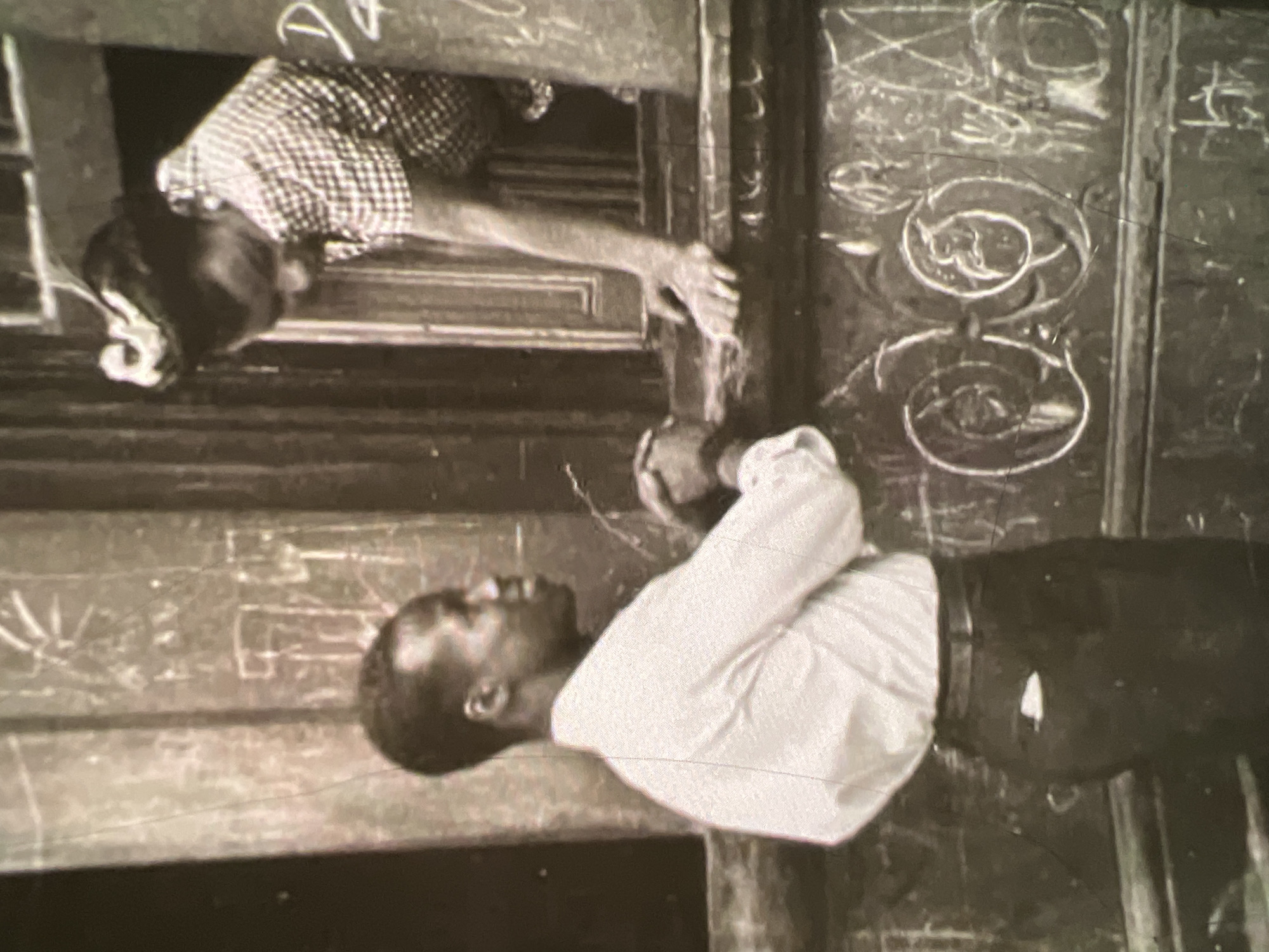 In a black and white photograph, a dark-skinned man stands looking up at a dark-skinned woman in a window. The man is wearing a light-colored collared shirt and dark trousers. The woman is wearing a checkered shirt and her hair is styled up. She is leaning out of the window, reaching down towards the man. The background shows a textured wall with what appears to be writing or drawings etched into it. The scene seems to be an outdoor urban setting, possibly in Lagos, Nigeria, given the architectural style and the people. The overall tone of the image is candid and intimate, capturing a moment of human interaction.