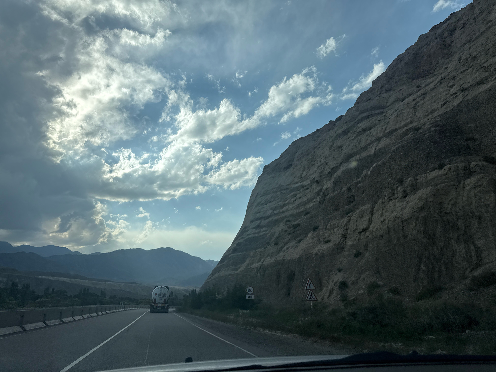 The image depicts a highway cutting through a mountainous landscape under a dramatic sky. A large, white tanker truck with a cylindrical tank and the letters "LPG" visible on its side is traveling on the right lane of the asphalt road. The road is bordered by concrete barriers on the left. To the right of the road, a massive, sheer rock face dominates the scene, showing layered geological formations and some sparse vegetation at its base. Several road signs are visible near the rock face: a circular sign with "90" inside, indicating a speed limit, and a triangular sign with a curved arrow indicating a sharp turn, followed by a pedestrian crossing sign. In the distance, rolling mountains form a hazy backdrop under a sky filled with a mix of dark and bright clouds, with sunlight breaking through in patches. The overall impression is of a remote, possibly arid, mountainous region.