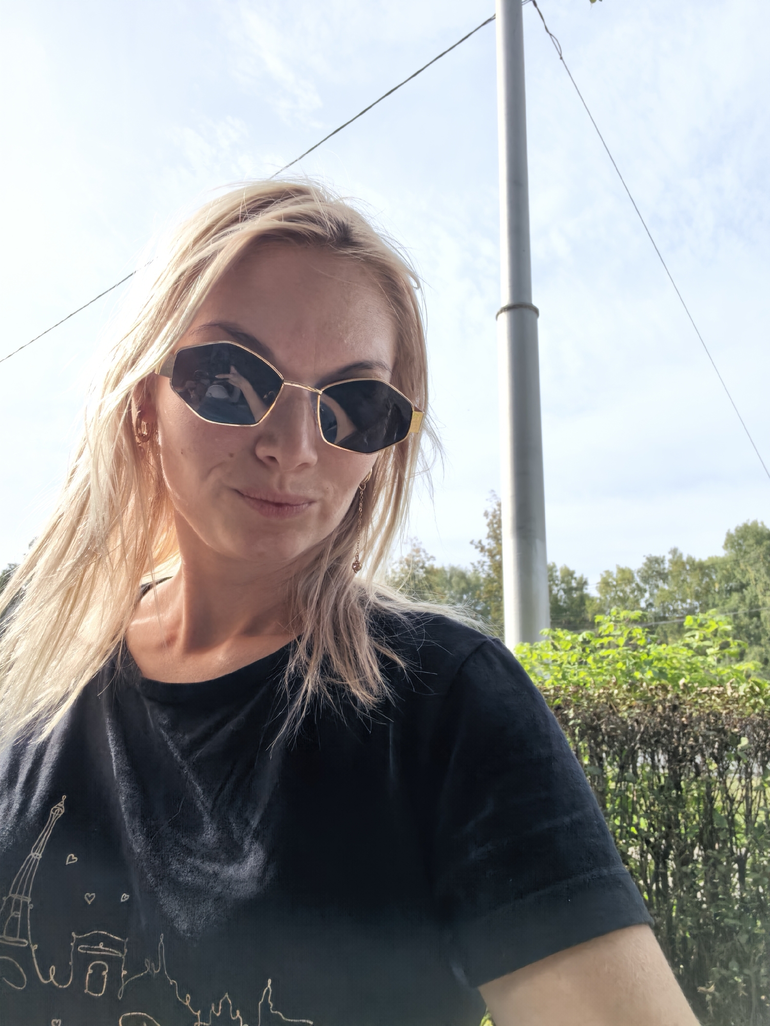 In an outdoor, daytime setting, a fair-skinned woman with blonde hair and sunglasses is captured in a close-up, selfie-style photograph. She is wearing a dark t-shirt with a graphic print depicting the Eiffel Tower and other Parisian landmarks. The background shows greenery and a pole, possibly a utility pole, against a sky with some clouds. The lighting suggests it is a sunny day. There is no indication that this scene is located in Sterlitamak, Russia.