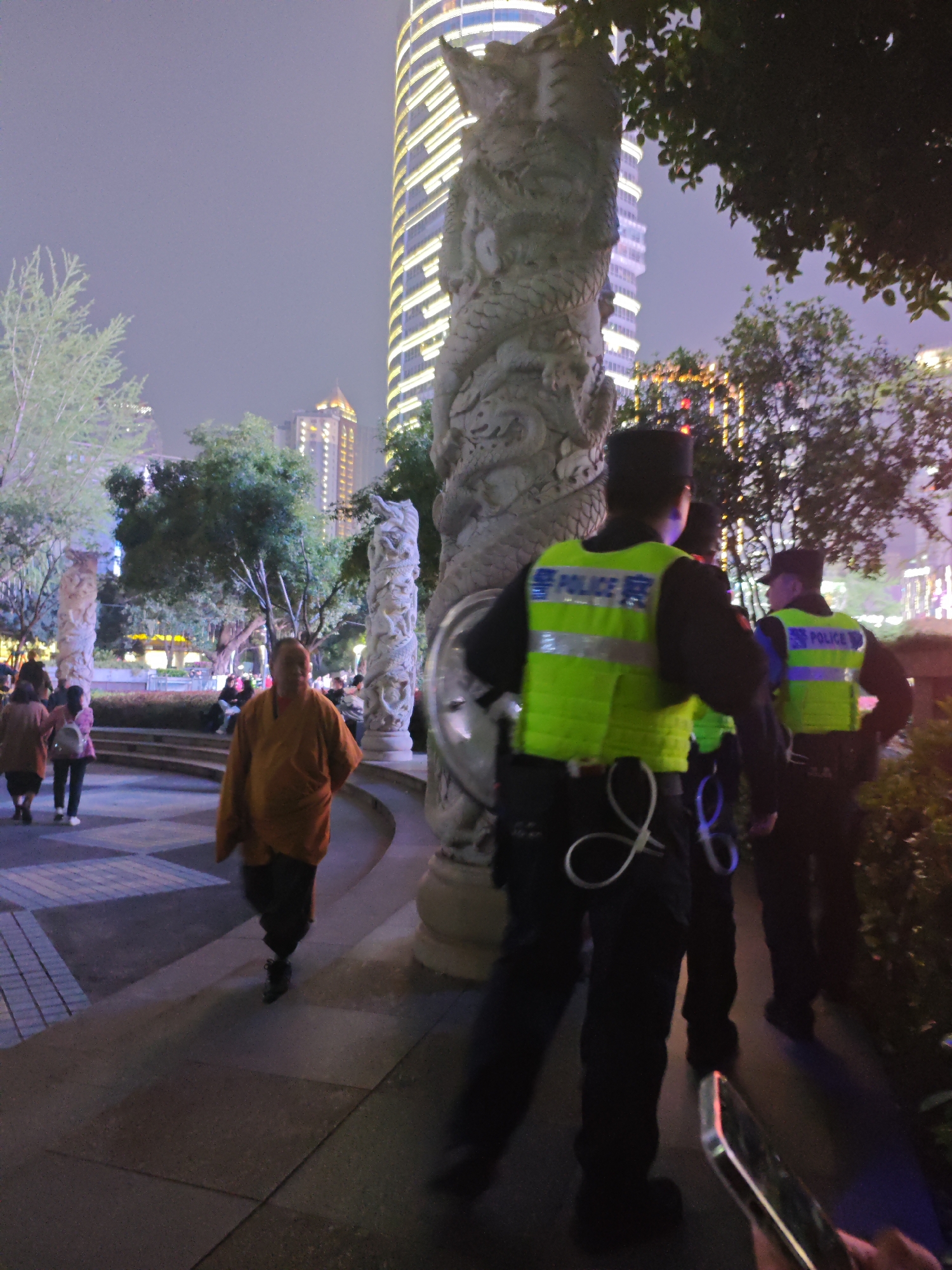 The image captures a scene in Liangjiang, China, at night. A monk dressed in orange robes walks on a paved path. Three uniformed police officers are standing on the path, with their backs to the camera. The police officers wear lime green vests with "POLICE 察" printed on the back. A tall, illuminated building stands in the background. To the left, there is a park area with trees and people. Decorative pillars with dragon carvings are also visible. A hand holding a smartphone is visible at the bottom right.