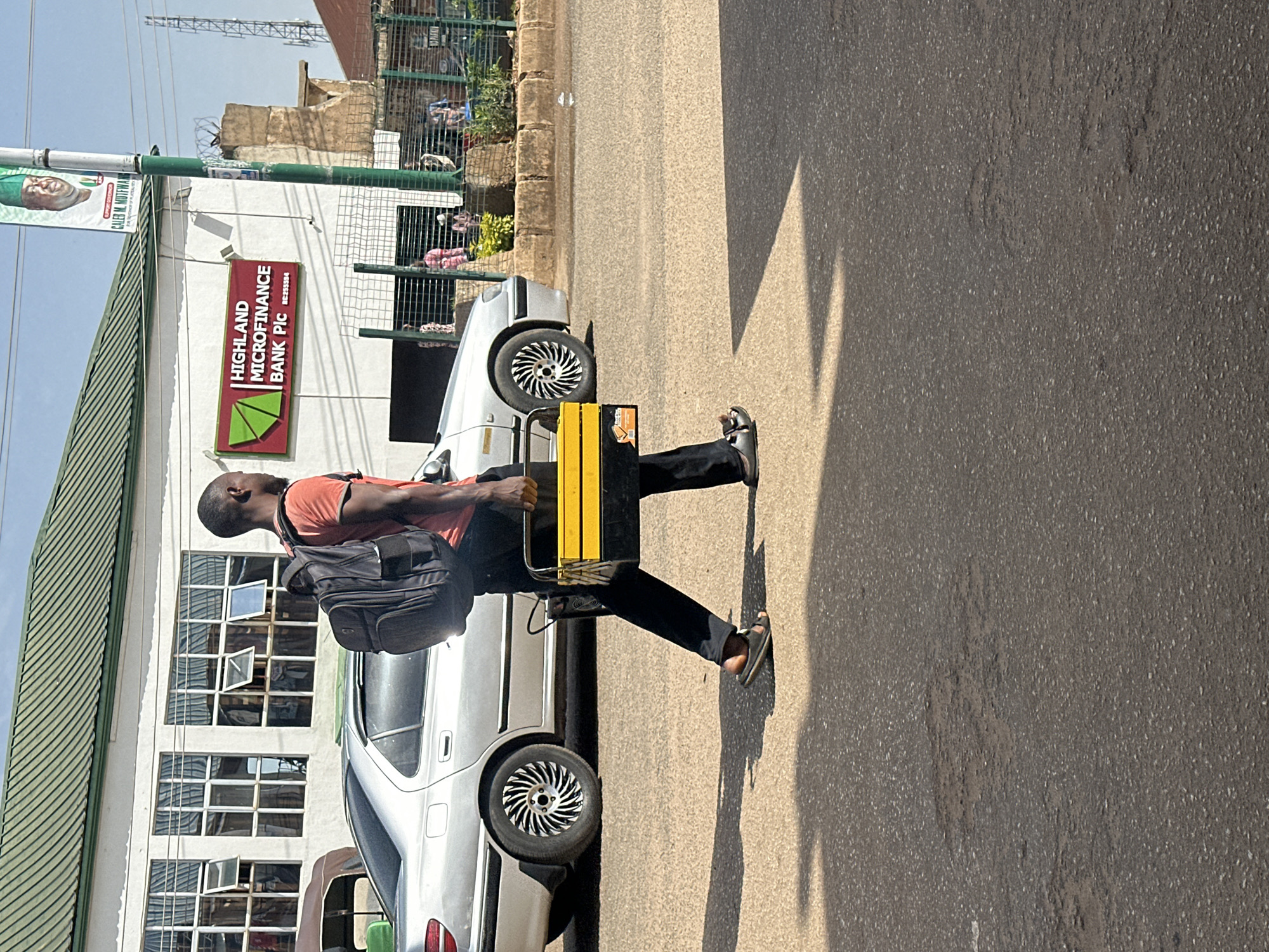 In Bukuru, Nigeria, a man walks on a paved road, carrying a black toolbox with yellow accents. He is wearing a red t-shirt, black pants, and sandals. A black backpack is on his back. Behind him, a silver sedan is parked. In the background, a white building with a green roof has the sign "HIGHLAND MICROFINANCE BANK PLC" with a logo. A banner with a picture of a man in a green hat hangs from a pole. The sky is clear and blue.