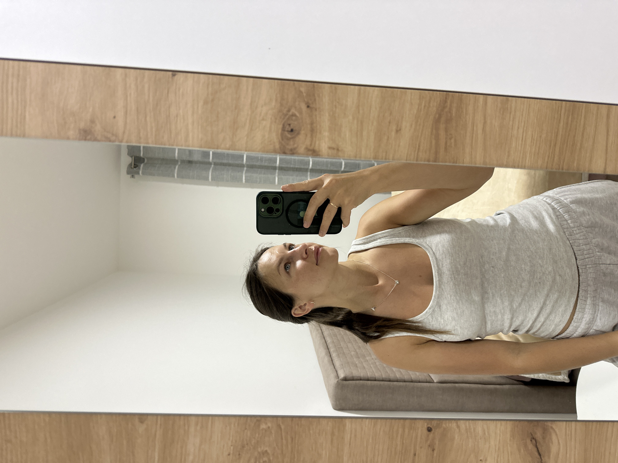 A woman takes a selfie in a mirror in an indoor setting, possibly a room in Donji Pobori, Montenegro. The room has white walls, light wood paneling, and a grey and white striped curtain in the background. The woman is wearing a grey tank top and grey shorts. She holds a black phone with a ring attachment. A bed with a light brown headboard is partially visible.