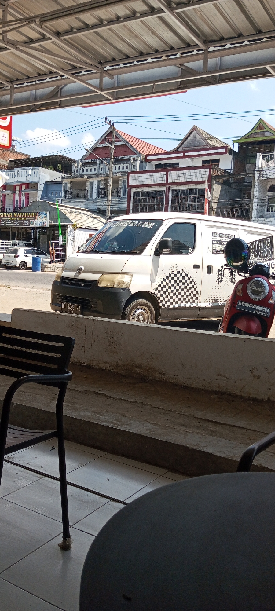 The image depicts a street scene in the city of Lempa, Indonesia. In the foreground, a black metal chair is visible, with a portion of a dark, round table to its right. A concrete barrier runs horizontally across the middle of the frame, separating the foreground from the street.
Behind the barrier, a white van is parked. The van has a checkered pattern on its side and text that appears to be in Indonesian. A red motorcycle with a helmet on its seat is parked to the right of the van. The license plate of the van reads "DD 8429 MB" and the motorcycle's license plate reads "DW 3552 MT".
In the background, buildings line the street, exhibiting a mix of architectural styles. The sky is clear with some clouds. The overall impression is a typical street view in a commercial area, likely during daylight hours given the illumination.