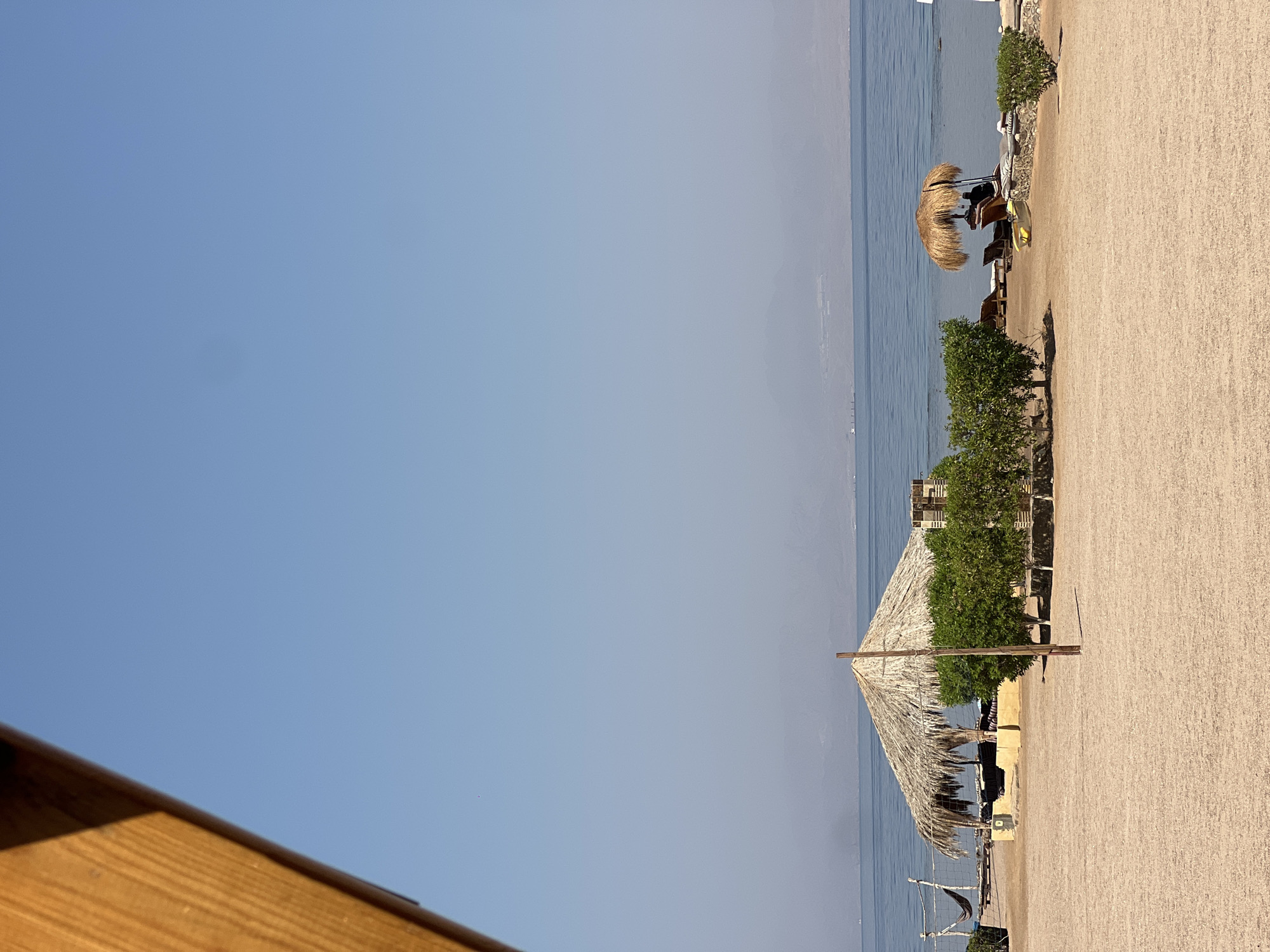 This is a medium-shot, eye-level, horizontal, outdoor photograph of a sandy beach in Nueiba, Egypt, on a clear, sunny day. The foreground is dominated by a wide expanse of tan-colored sand. In the mid-ground, a large thatched-roof hut is situated on the sand, with a volleyball net and hammock visible behind it to the left. A small cluster of green trees separates the hut from a second, smaller thatched-roof umbrella that shelters a lounge chair and a person. In the background, the calm, blue sea stretches to the horizon, with a hazy, mountainous coastline visible in the distance. The sky is a pale, clear blue. A wooden beam from a structure is visible in the upper left corner of the frame, casting a shadow over part of the sky.