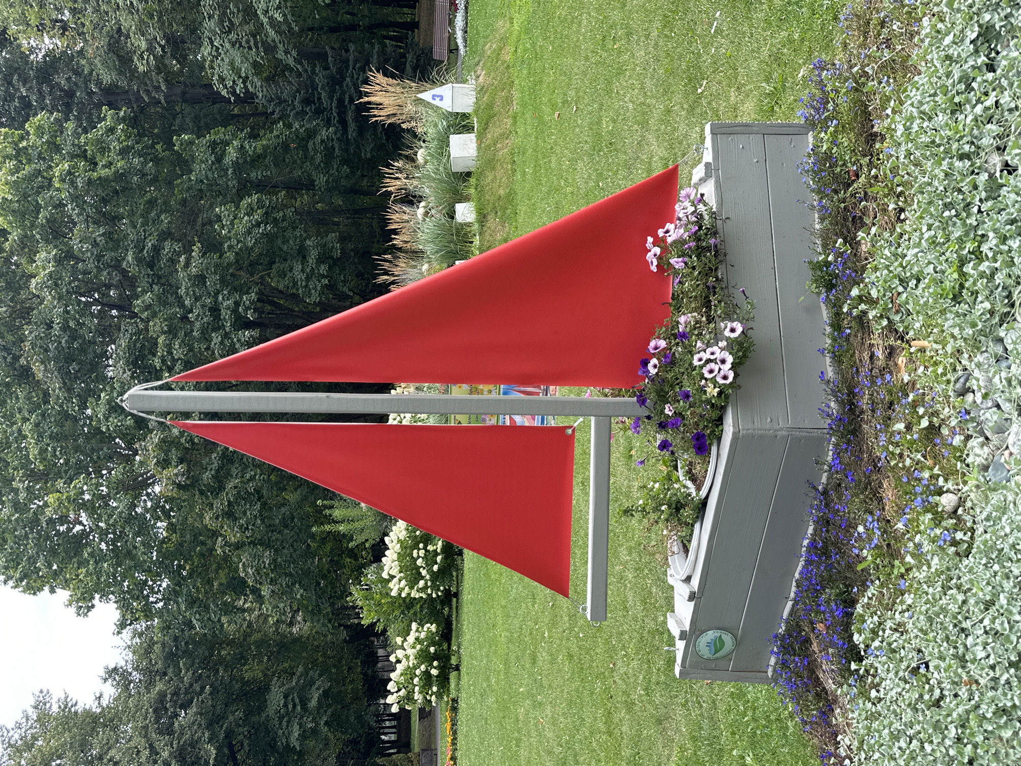 This is a forensic, matter-of-fact description of an image taken in Minsk, Belarus. The image shows a garden decoration in the shape of a sailboat with red sails, positioned in a park. The sailboat is made of a light gray wooden box acting as the hull, filled with flowers, including purple and white petunias, and bordered by small blue flowers. Behind the sailboat, lush green grass covers the ground, with a variety of trees and bushes forming a dense backdrop. In the distance, a park bench is visible, along with ornamental grasses and white cube-shaped structures. The sky is overcast. The overall scene depicts a well-maintained public green space.