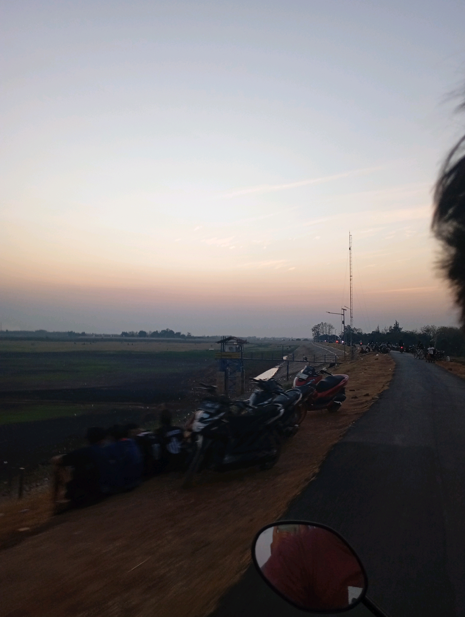 The image depicts a view from a moving vehicle, likely a motorbike given the presence of a side mirror in the foreground and the perspective. It is evening in Haurkolot, Indonesia. The scene shows a road curving to the right, with several motorbikes parked along its edge. On the left side of the road, a group of people is seated on a grassy embankment, facing away from the camera. Beyond the embankment, a vast, flat agricultural field stretches towards the horizon, with scattered trees and buildings visible in the distance. The sky is a gradient of soft blues and oranges, indicative of sunset or sunrise. A tall communication tower stands prominently on the right side of the road, further into the distance.