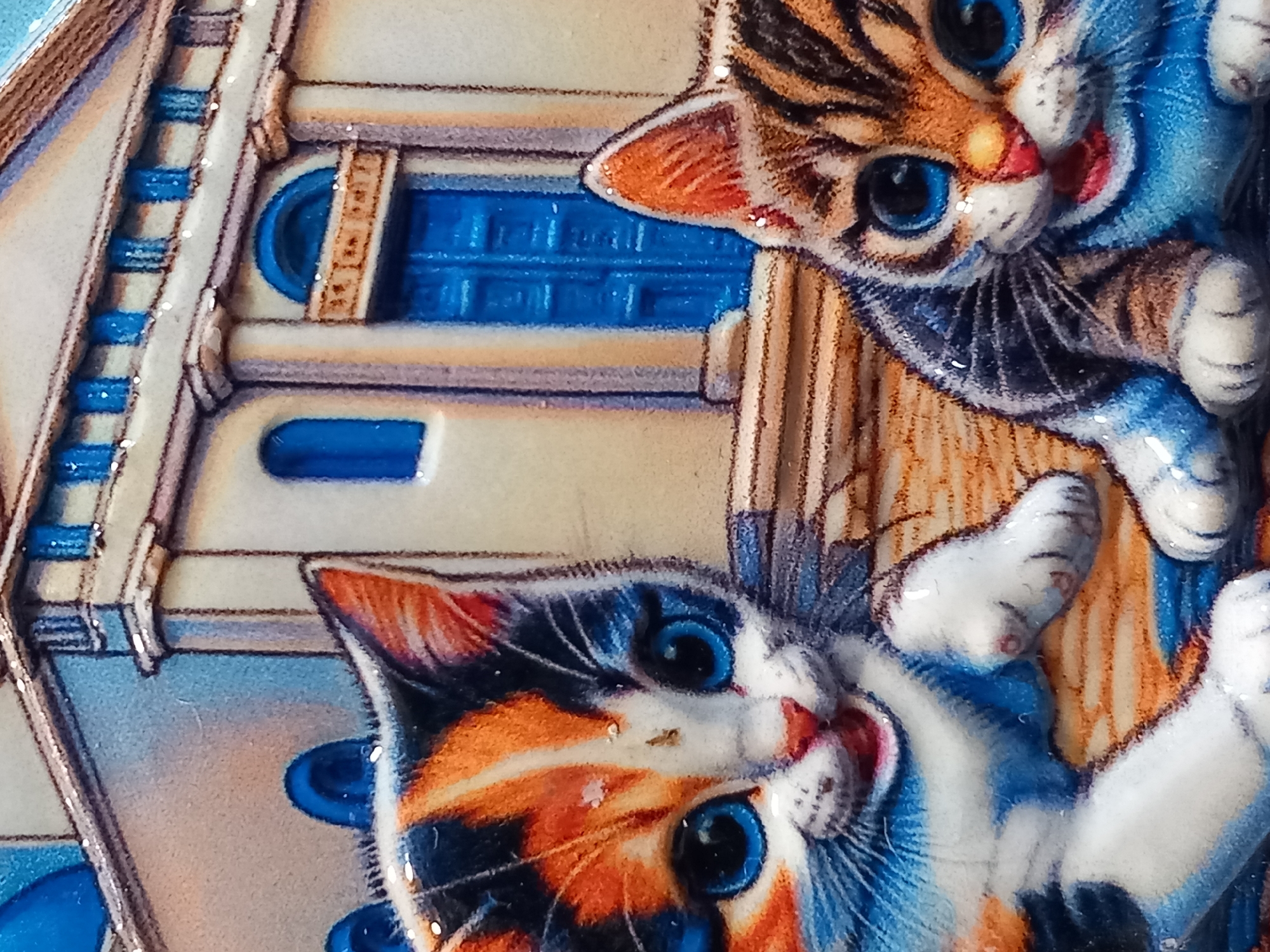 This is a close-up, high-angle photograph of a decorative item featuring two kittens rendered in a raised, embossed style. The kittens are depicted with bright blue eyes, intricate fur patterns in orange, black, and white, and rosy cheeks. One kitten, in the foreground, has its head tilted and its mouth slightly open, giving it a playful or surprised expression. The other kitten, partially behind and above the first, also has large blue eyes and is looking towards the viewer.
The background appears to be a stylized building or structure with light-colored walls and bright blue architectural details, possibly windows or doors, with ribbed patterns. The overall impression is one of a charming, whimsical scene, likely part of a collectible item or artwork. The lighting is bright, highlighting the glossy texture and raised details of the figures. There is no visible text in the image. The context of the location and any specific event is unknown.