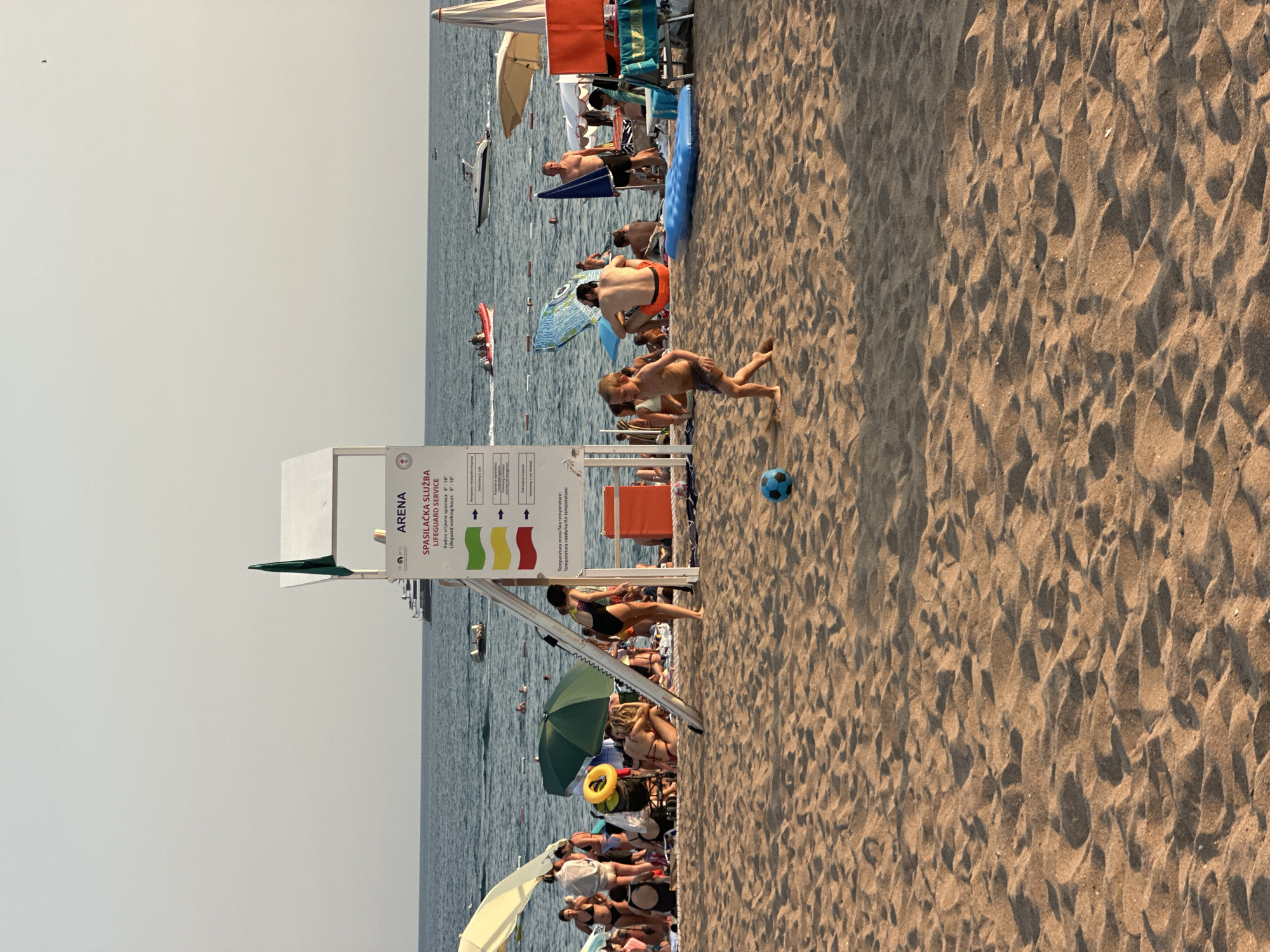 In Boreti, Montenegro, a sandy beach is populated with numerous beachgoers enjoying the sunny weather. A young boy is actively playing with a blue soccer ball in the foreground. Beach umbrellas and towels dot the sand, providing shade for individuals and groups lounging. In the background, the blue sea stretches to the horizon, dotted with several boats, including a white speedboat and a smaller red boat with passengers. A lifeguard tower stands prominently on the beach, displaying information about lifeguard services and sea temperature. The overall scene captures a lively and relaxing atmosphere at the beach.