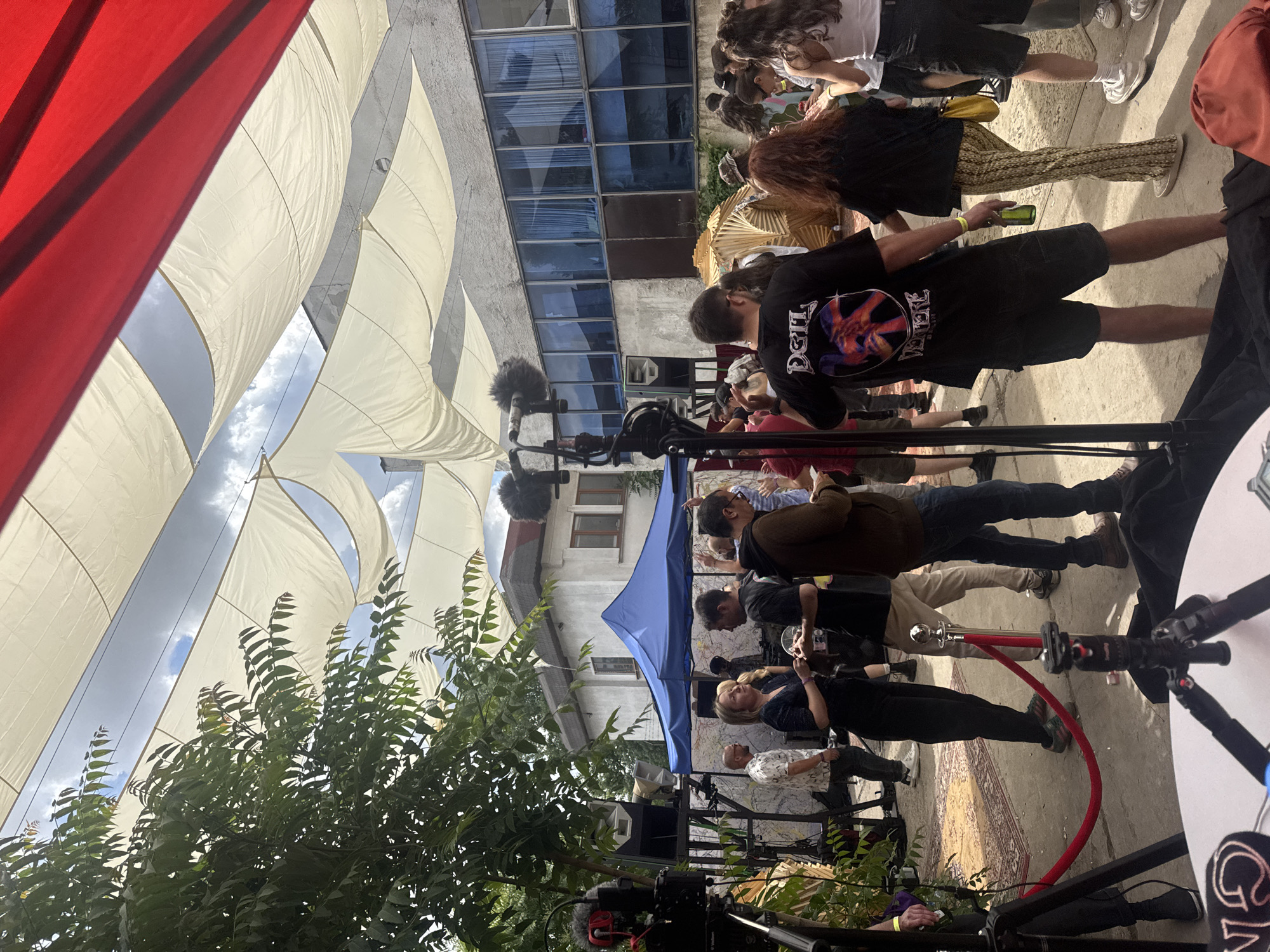 The image depicts an outdoor gathering in Sofia, Bulgaria, likely a festival or event. The scene is characterized by a casual atmosphere with a diverse group of people. Overhead, several white fabric canopies are stretched, creating a shaded area, with a red canopy visible at the top right. In the background, a building with large blue-tinted windows is present.
Several individuals are captured in various states of interaction. Some are standing and conversing, while others appear to be observing or moving through the space. A blue tent is set up, possibly serving as a vendor or information booth. There is a red rope barrier on the ground, suggesting an entrance or a designated area.
A prominent feature in the foreground is camera equipment, including tripods and microphones, indicating that the event is being filmed or recorded. Various people are dressed in casual attire, reflecting the relaxed nature of the gathering. The ground surface appears to be concrete, with a patterned rug visible in the middle ground. The presence of trees and foliage suggests an outdoor setting within a potentially urban environment. The overall impression is one of a vibrant social event with a touch of professional documentation.
