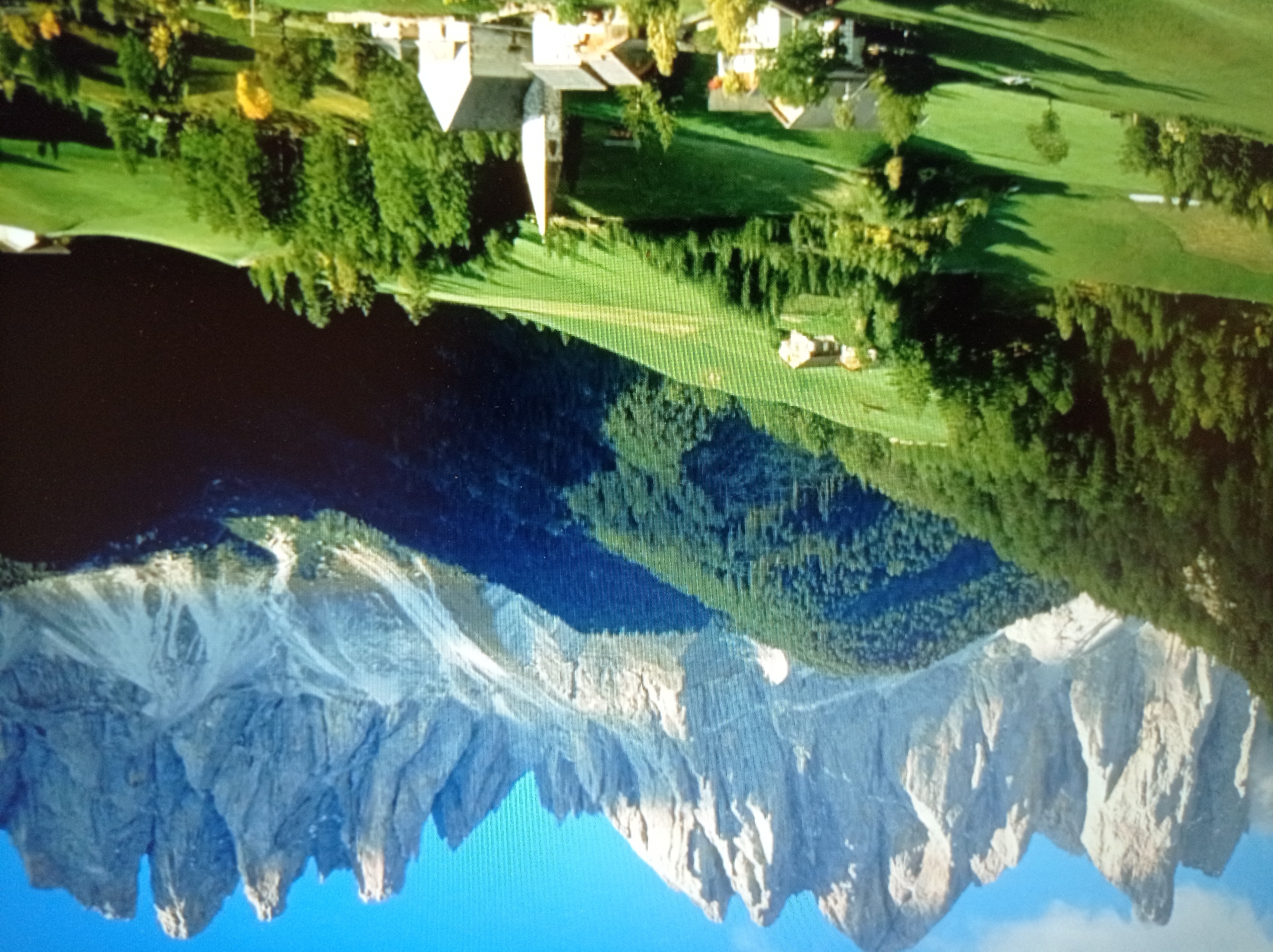 This is a photograph of a scenic mountain landscape with a small village in the foreground. The jagged, rocky peaks of the mountains dominate the background, with patches of snow visible on their upper slopes. The midground features lush green hills covered in dense pine forests. In the foreground, a charming village is nestled amongst rolling green meadows. Several buildings, including houses and a church with a tall spire, are visible. The sun is shining, casting shadows across the landscape and highlighting the vibrant green of the fields and trees. There are no people visible in the image. The scene depicts a serene and picturesque rural setting.