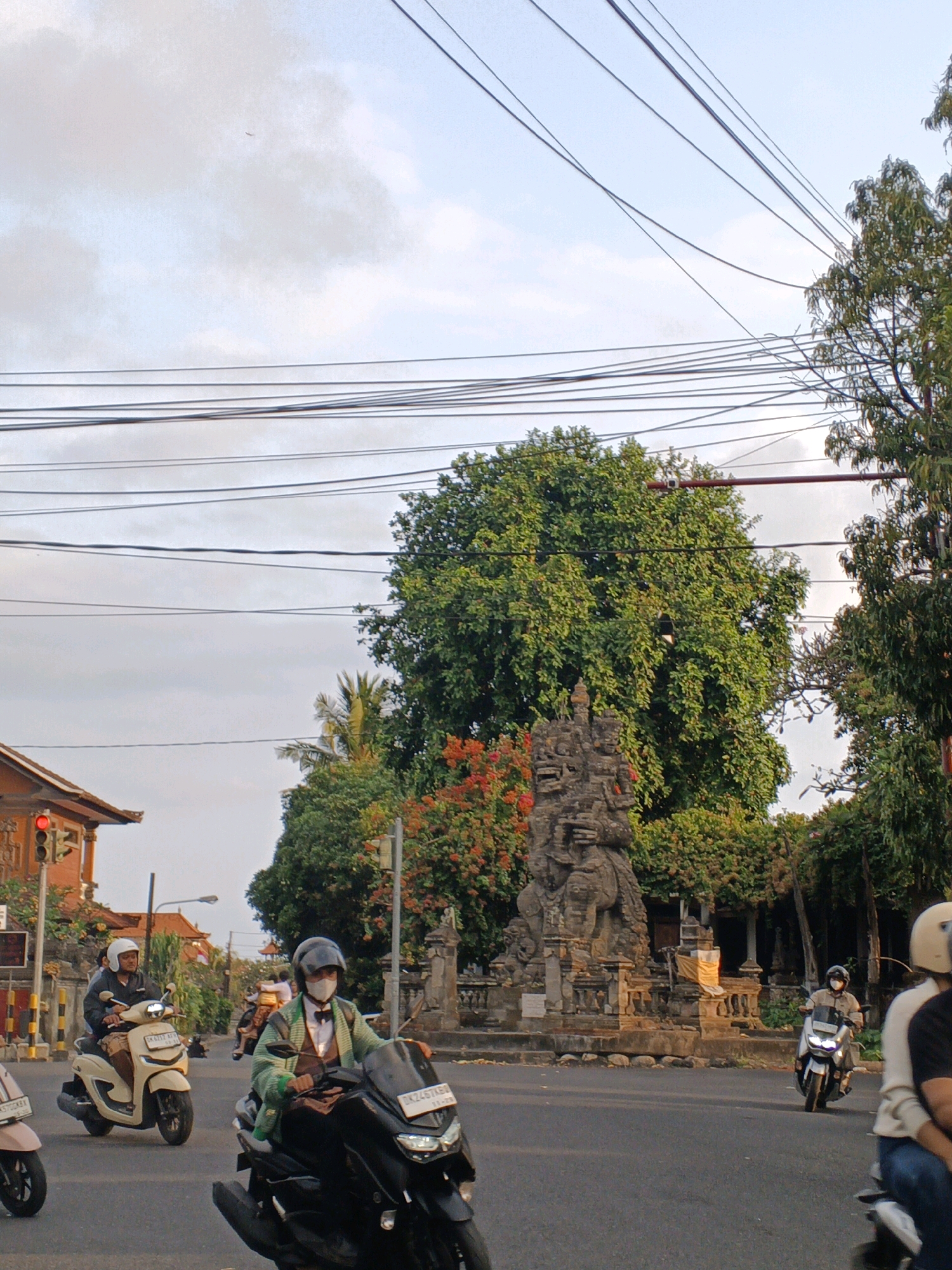 In Sukawati, Indonesia, a traffic scene unfolds under an overcast sky, with power lines crisscrossing overhead. A light-colored scooter, driven by a person wearing a white helmet, is in the foreground on the right, accompanied by a person in the back seat also wearing a white helmet. To the left, a black scooter is ridden by a person wearing a black helmet and a face mask, dressed in a green cardigan over a brown collared shirt with a bow tie. Behind them, a light-colored scooter is operated by a person with a white helmet. In the background, a stone monument stands prominently, surrounded by lush green trees and vibrant red flowers. To the left, a building with a red-tiled roof and a traffic light showing red is visible. The road is paved, and the overall impression is that of a busy street with traditional Balinese architectural elements.