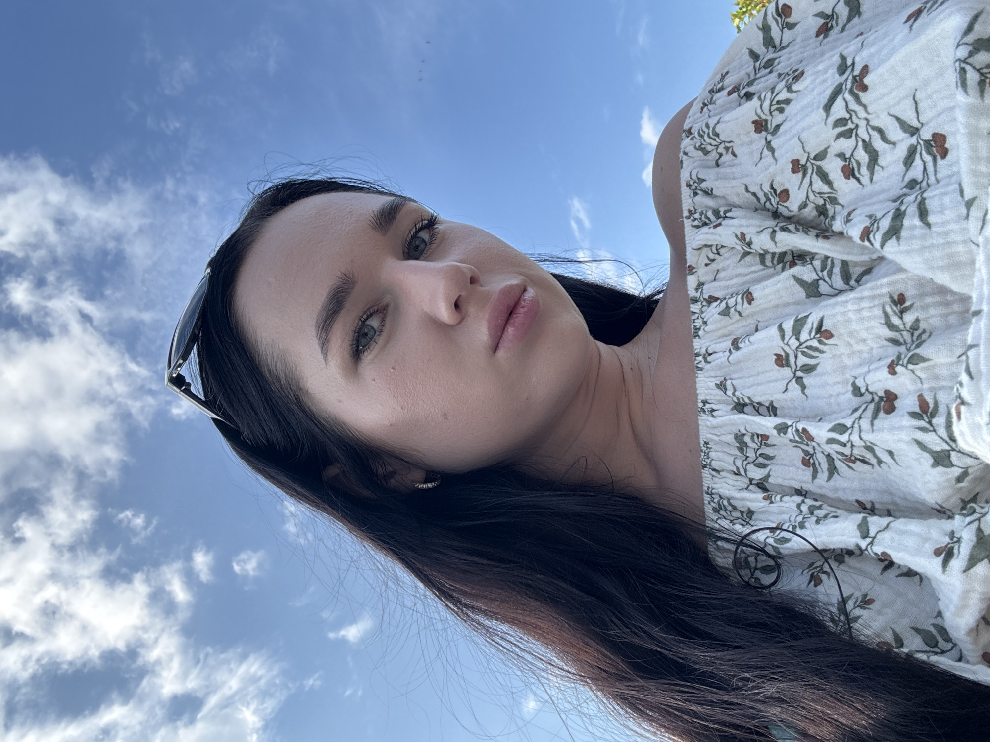 The image shows a close-up, low-angle shot of a young Caucasian woman outdoors against a blue sky with white clouds. She has long, dark hair and is wearing a white off-the-shoulder top with a floral pattern. Sunglasses are perched on top of her head. The woman's gaze is directed towards the camera with a neutral expression. The scene is entirely devoid of any identifiable landmarks or features that would suggest a specific location within Novosibirsk, Russia, or any other city.
