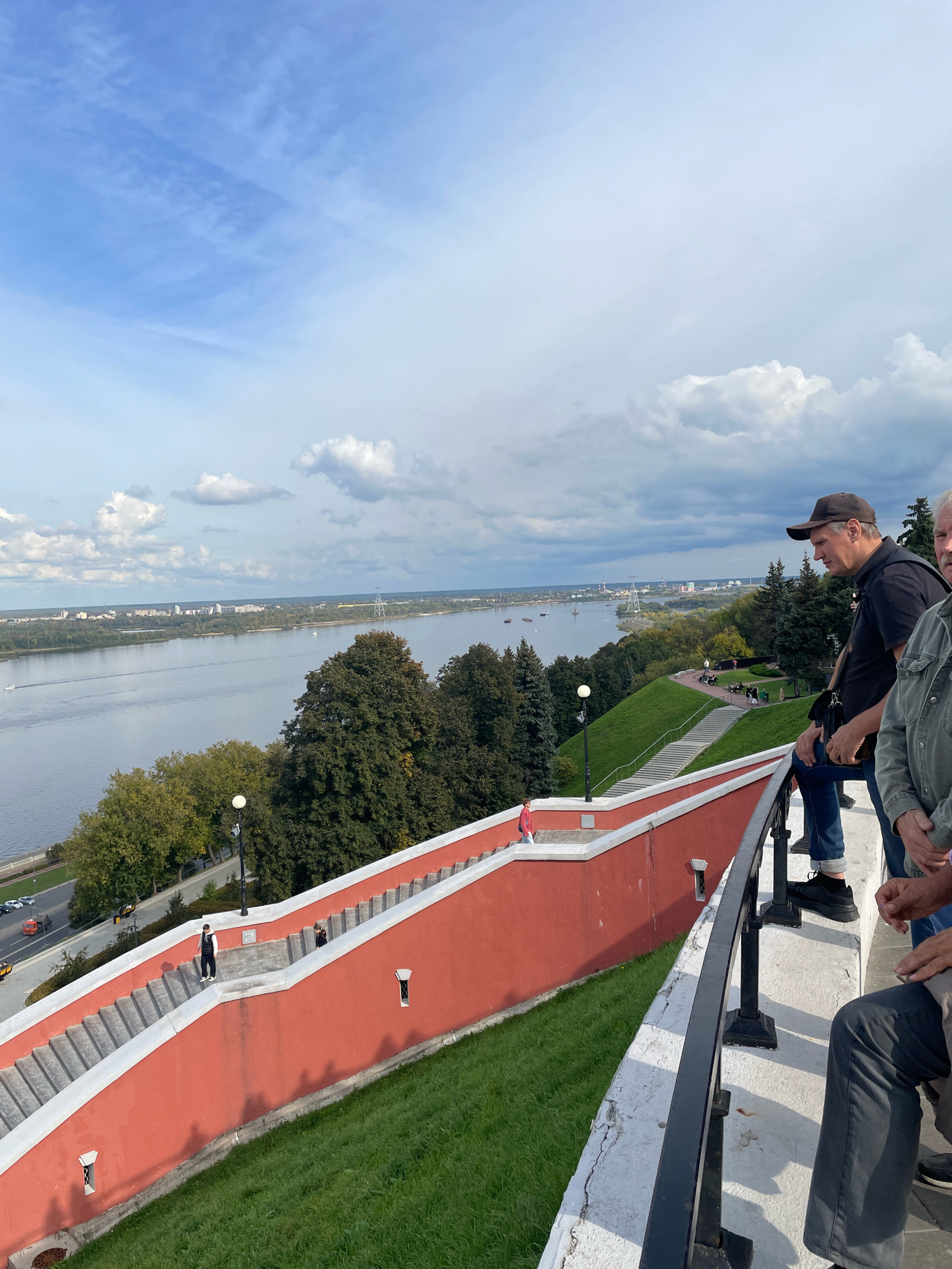 The image shows a wide river with a city skyline on the opposite bank. In the foreground, a red-walled staircase descends a grassy slope, with a black metal railing along the top. Several people are visible on the stairs and standing near the railing. One man in a black shirt and baseball cap is looking out over the river. This is a view of the Volga River from the Nizhny Novgorod Kremlin in Nizhny Novgorod, Russia. The sky is partly cloudy.