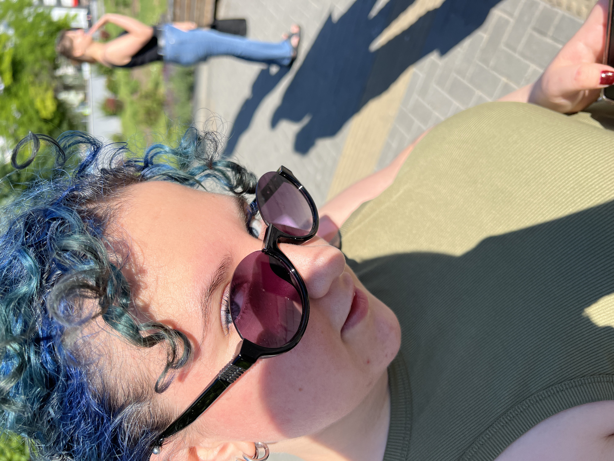 A young woman with blue curly hair and sunglasses looks down at her phone. She is wearing a green sleeveless top. In the background, a woman in a black tank top and blue jeans walks away from the camera. The scene appears to be an outdoor public space, possibly a park or plaza, with paving stones and trees visible. The image was taken in Chișinău, Moldova.