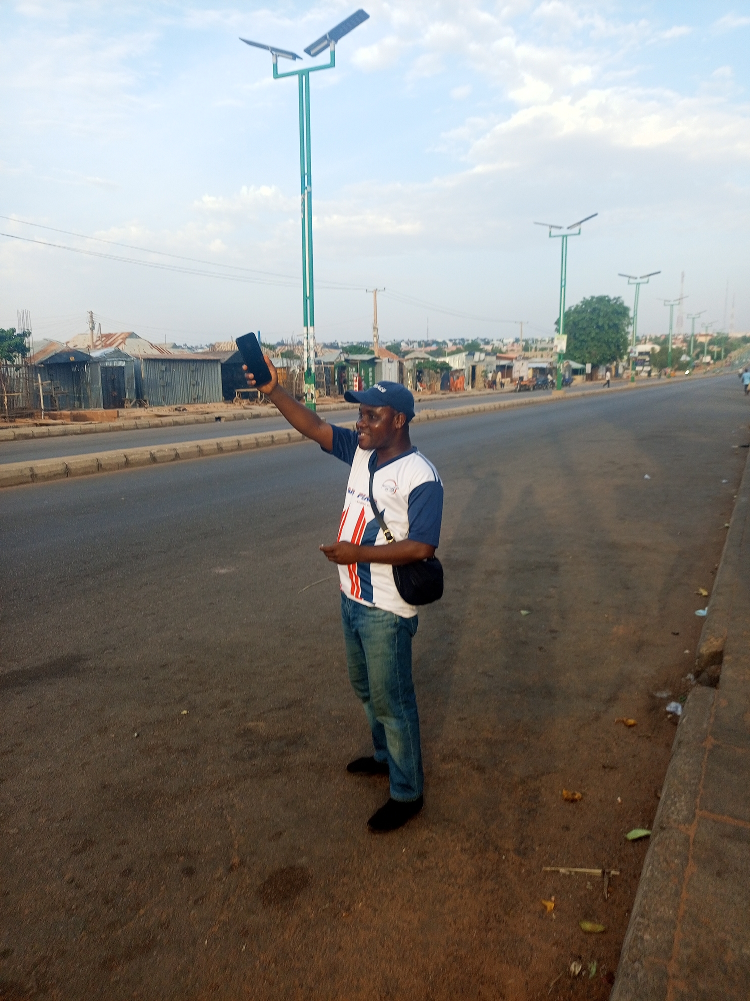 A man stands on the side of a road in what appears to be Tumfure, Nigeria. He wears a baseball cap, a white t-shirt with red and blue accents, blue jeans, and black shoes. He holds a black mobile phone in his raised right hand. He has a black bag slung across his shoulder. Behind him is a wide asphalt road with a median. Buildings of varying construction line the road, and green street lights stand tall. The sky is partly cloudy.